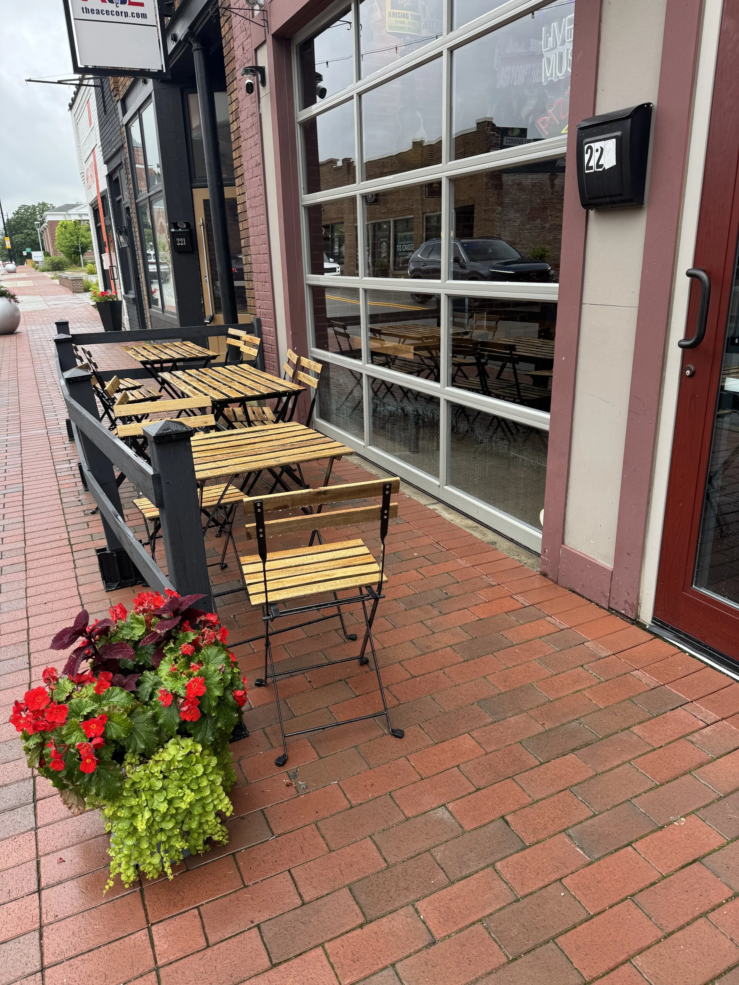 Empty outdoor seating area with wooden tables and chairs, potted plants, and a glass storefront on a brick sidewalk.