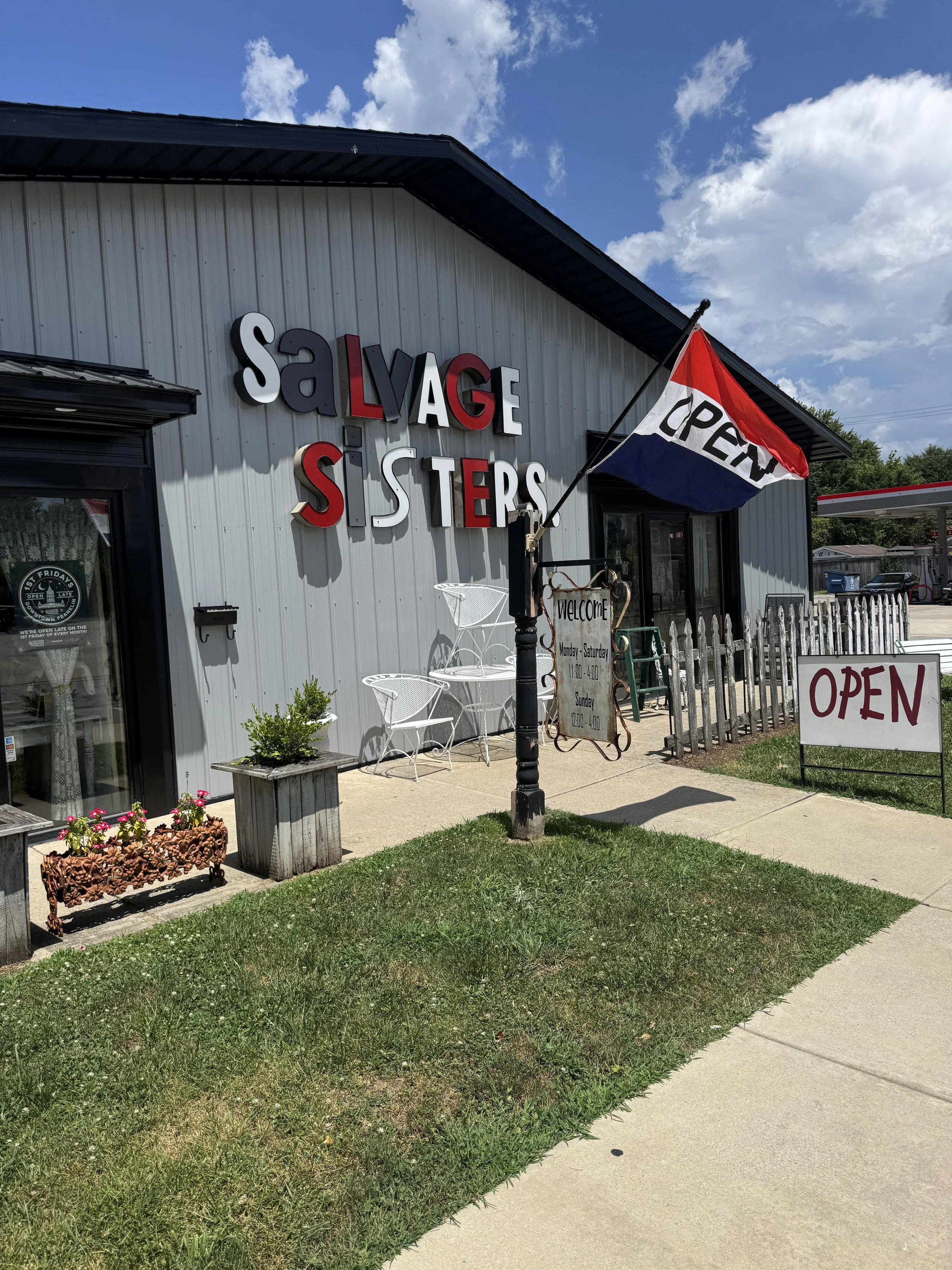 Exterior of a store with large sign reading 'Salvage Sisters,' an American flag, outdoor seating, and a white picket fence. The store has a welcome sign with hours. The sky is partly cloudy.