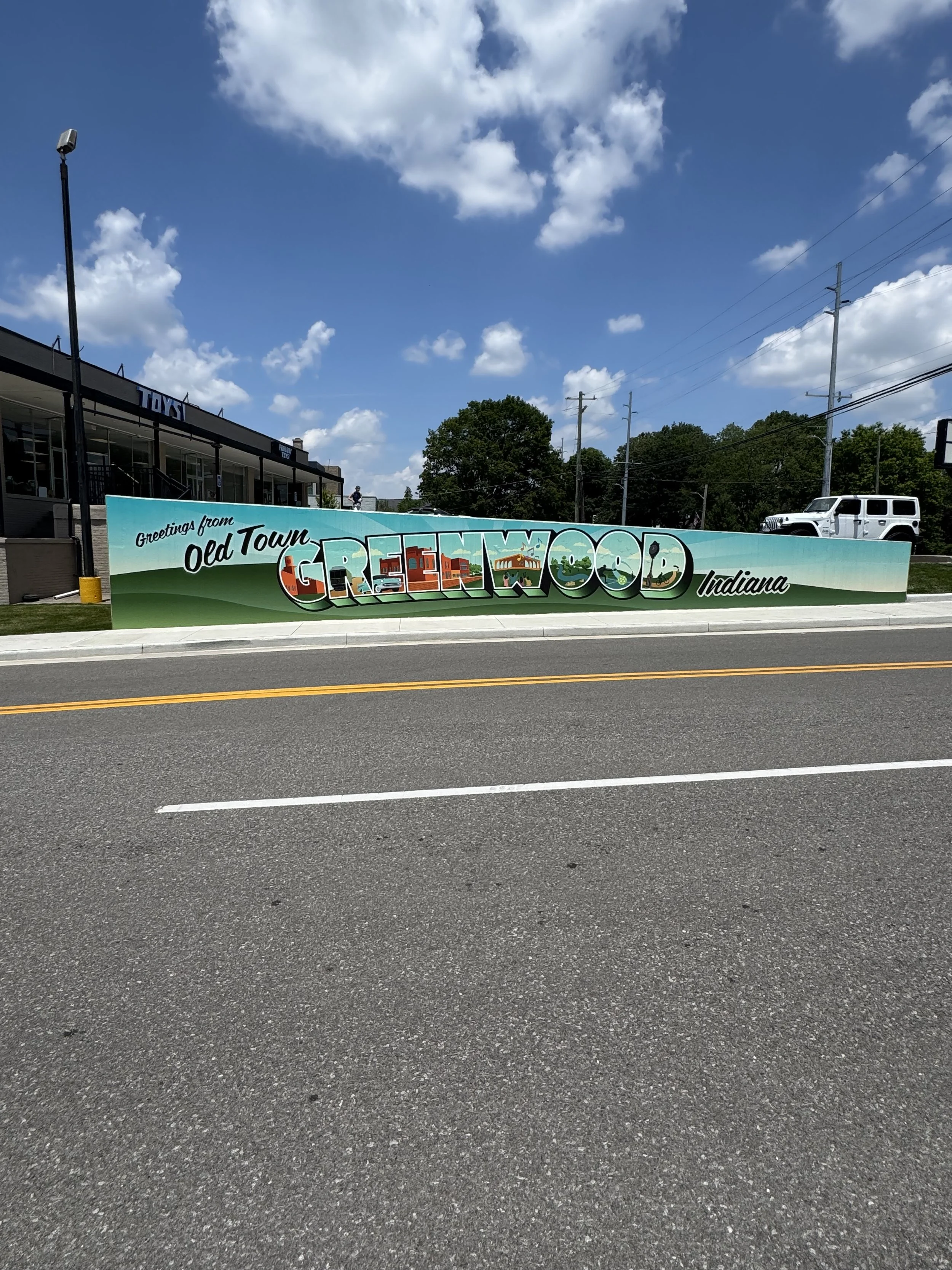 A colorful mural on a wall in Old Town, Greenwood, Indiana, greeting visitors with the words 'Greetings from Old Town, Greenwood Indiana' and illustrations of local landmarks and scenery, under a partly cloudy sky.