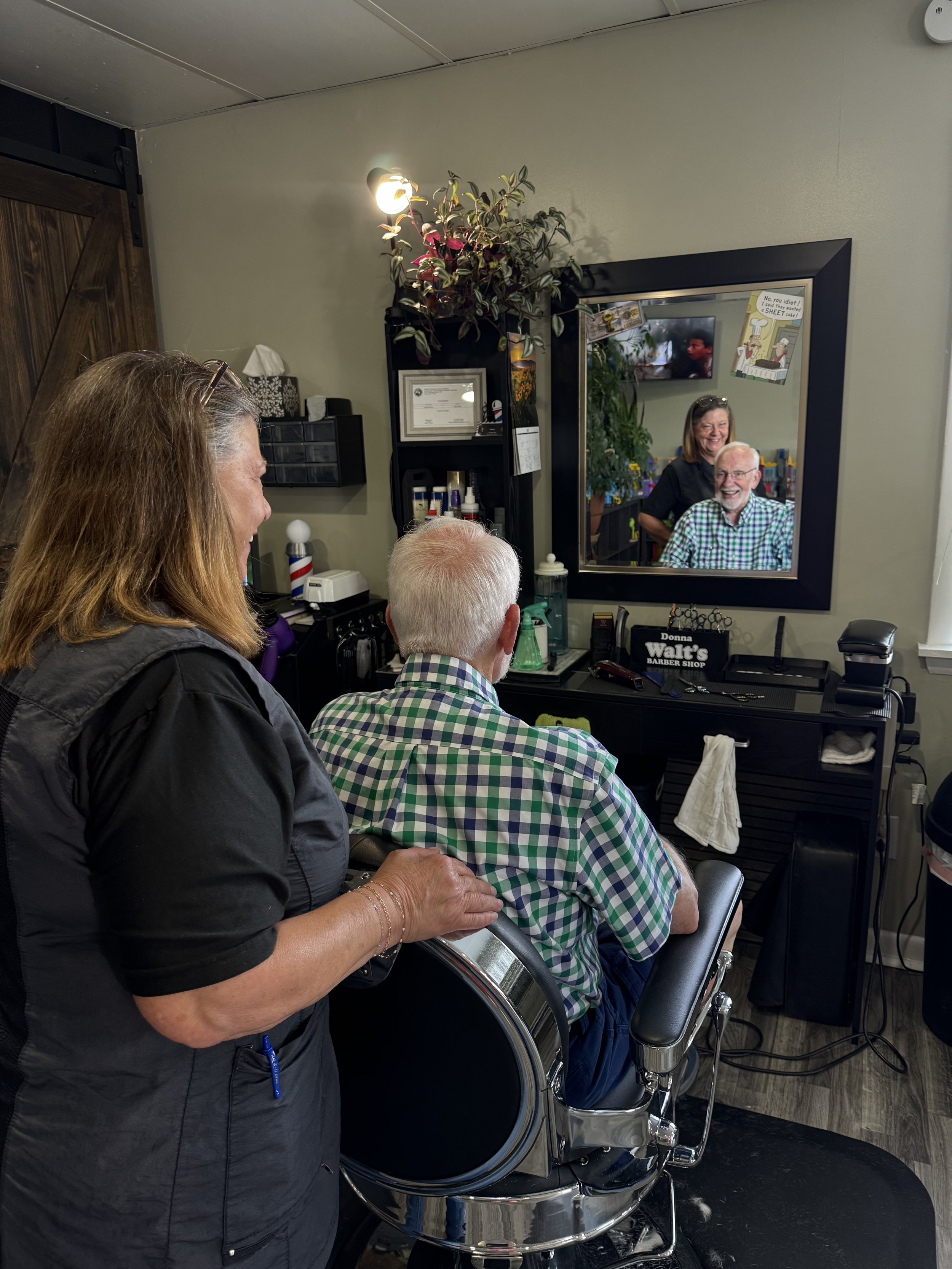 A man with white hair and glasses sitting in a barber chair, smiling, wearing a green and white checkered shirt, in a barbershop. Two women are standing nearby, one with long hair and a dark jacket, the other with short hair and glasses on her head, both smiling and looking at the man. The barbershop has a mirror, various products, and a TV on the wall.