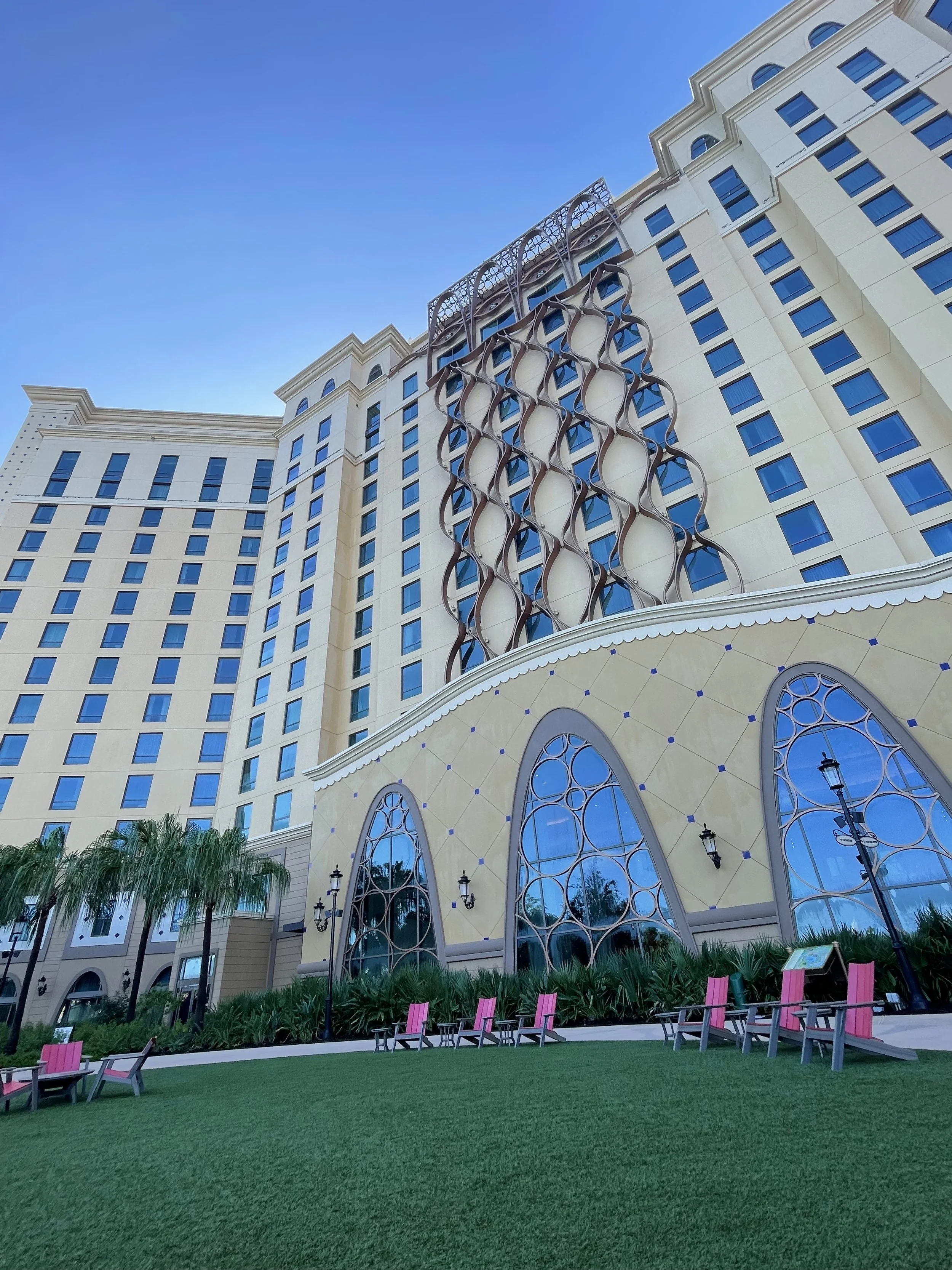 The exterior of a tall hotel building with a modern decorative design on its facade, set against a clear blue sky. There are pink and black benches on a well-maintained grass lawn in the foreground, along with lamp posts and some trimmed bushes.