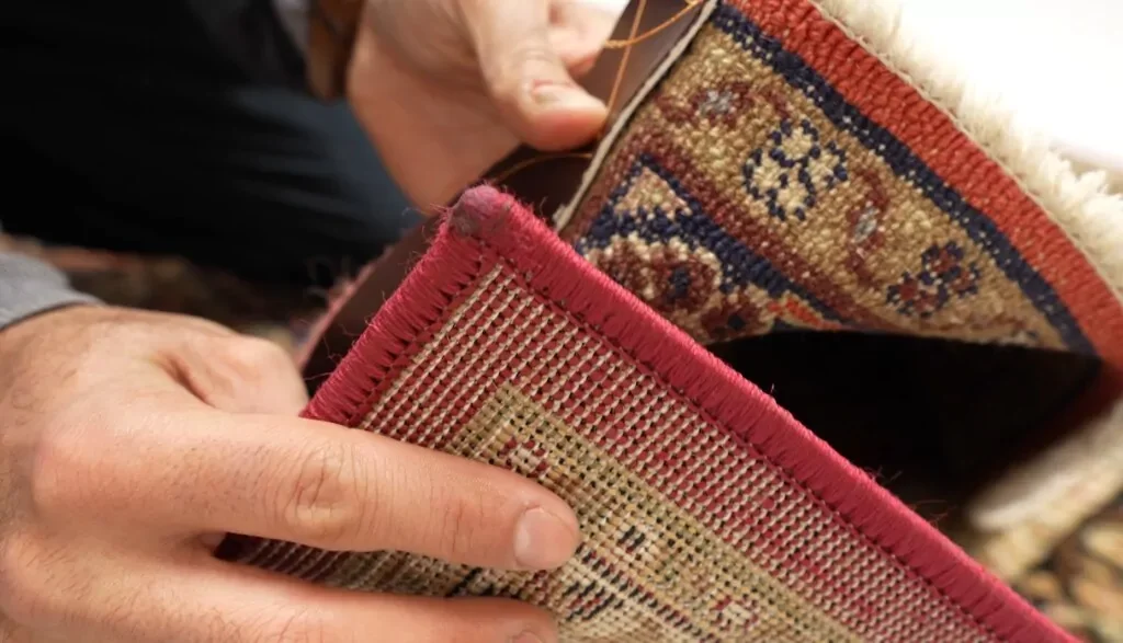Close-up of hands examining a small, rolled-up ornate rug with detailed patterns and a color palette including red, blue, and beige.