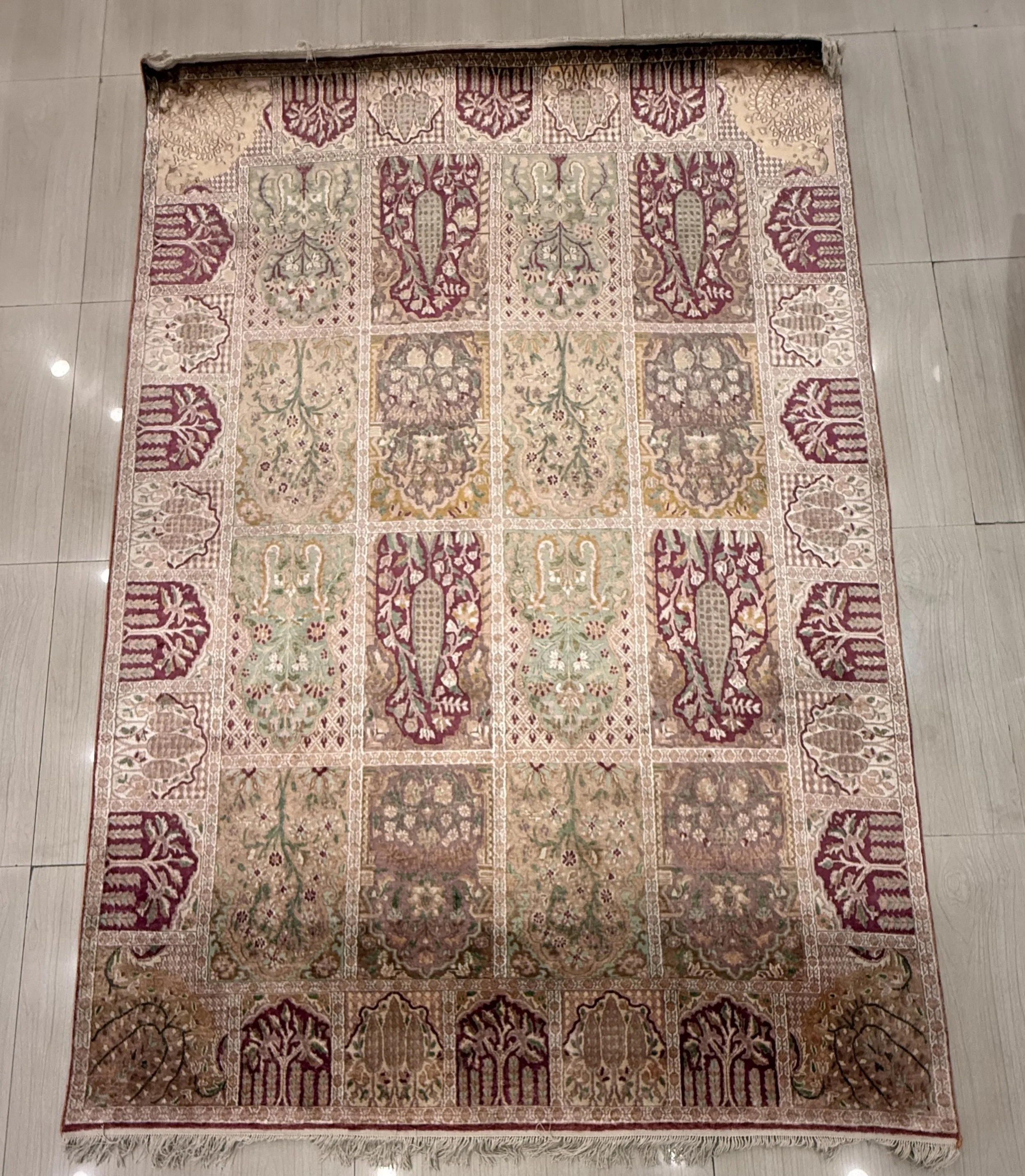 A rectangular area rug with intricate patterns and a border in shades of cream, red, green, and beige, placed on a light-colored tiled floor. The rug features a repeating floral and geometric design.