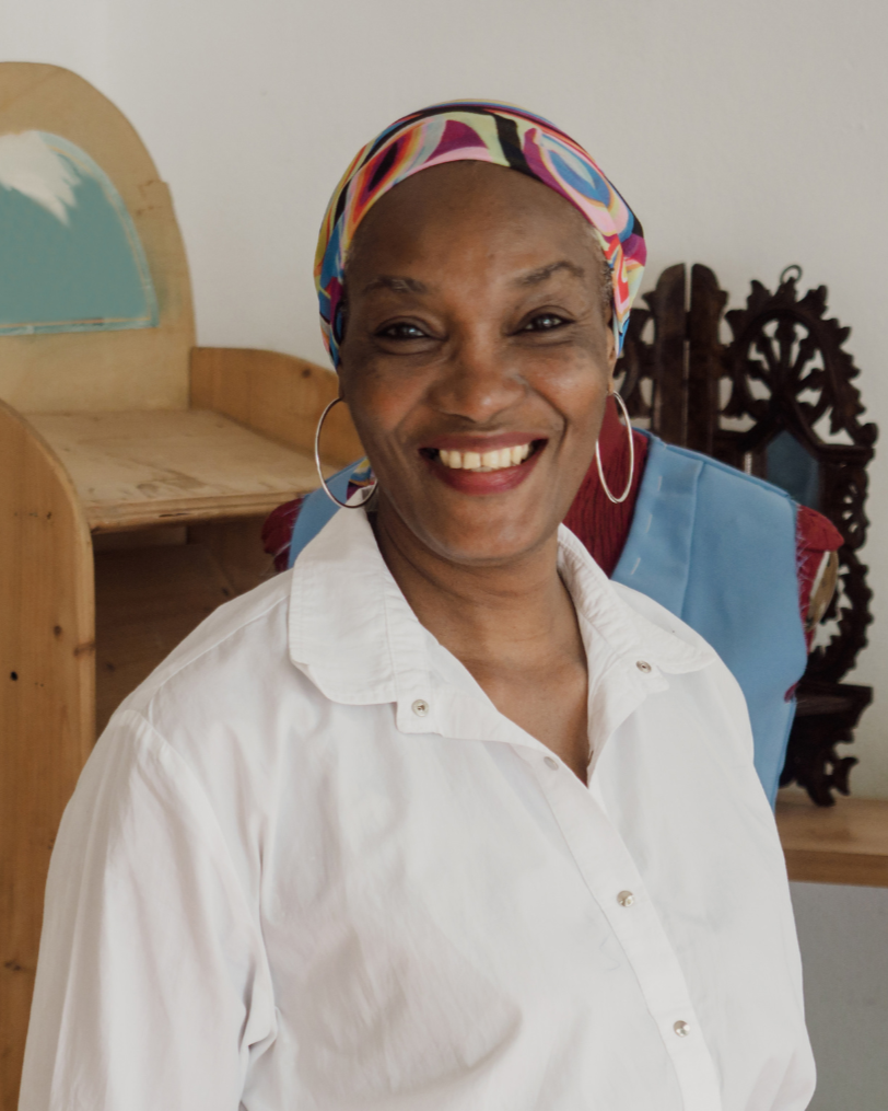 A smiling woman wearing a colorful headscarf, hoop earrings, and a white button-up shirt.