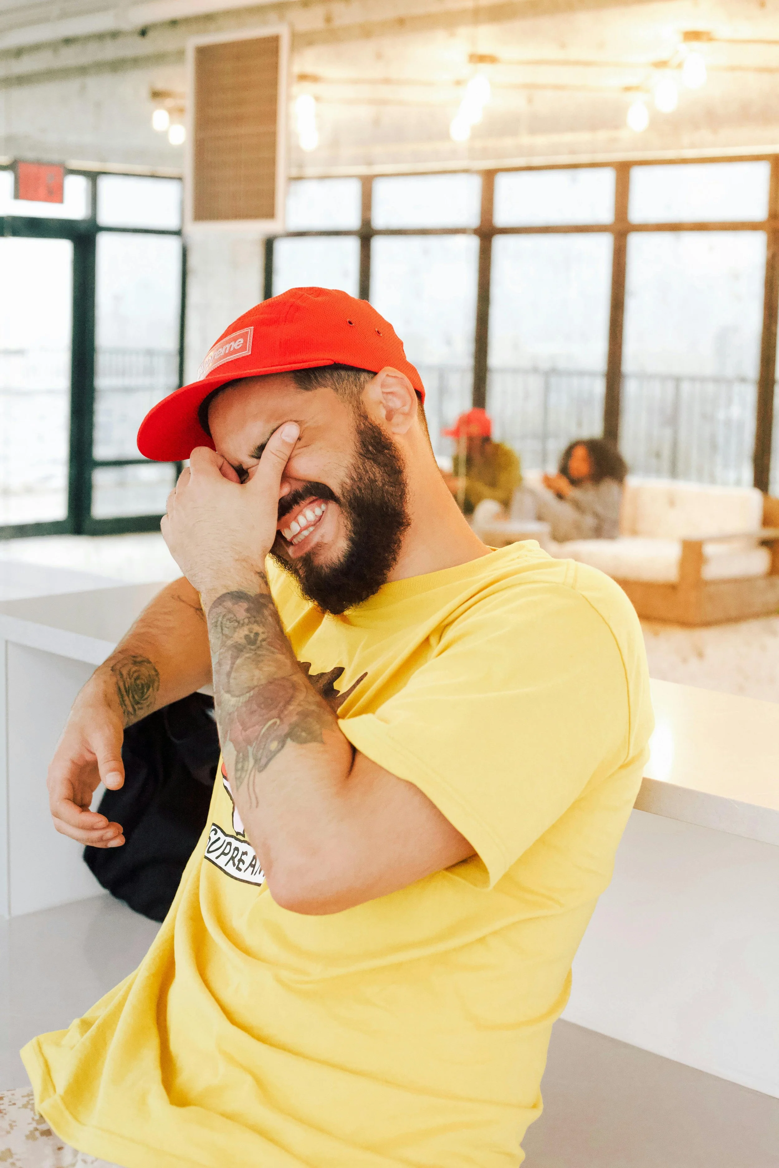 A man with a beard wearing a yellow T-shirt and a red cap is sitting and laughing with his hand covering part of his face in a bright indoor setting, with a woman in the background sitting on a couch.