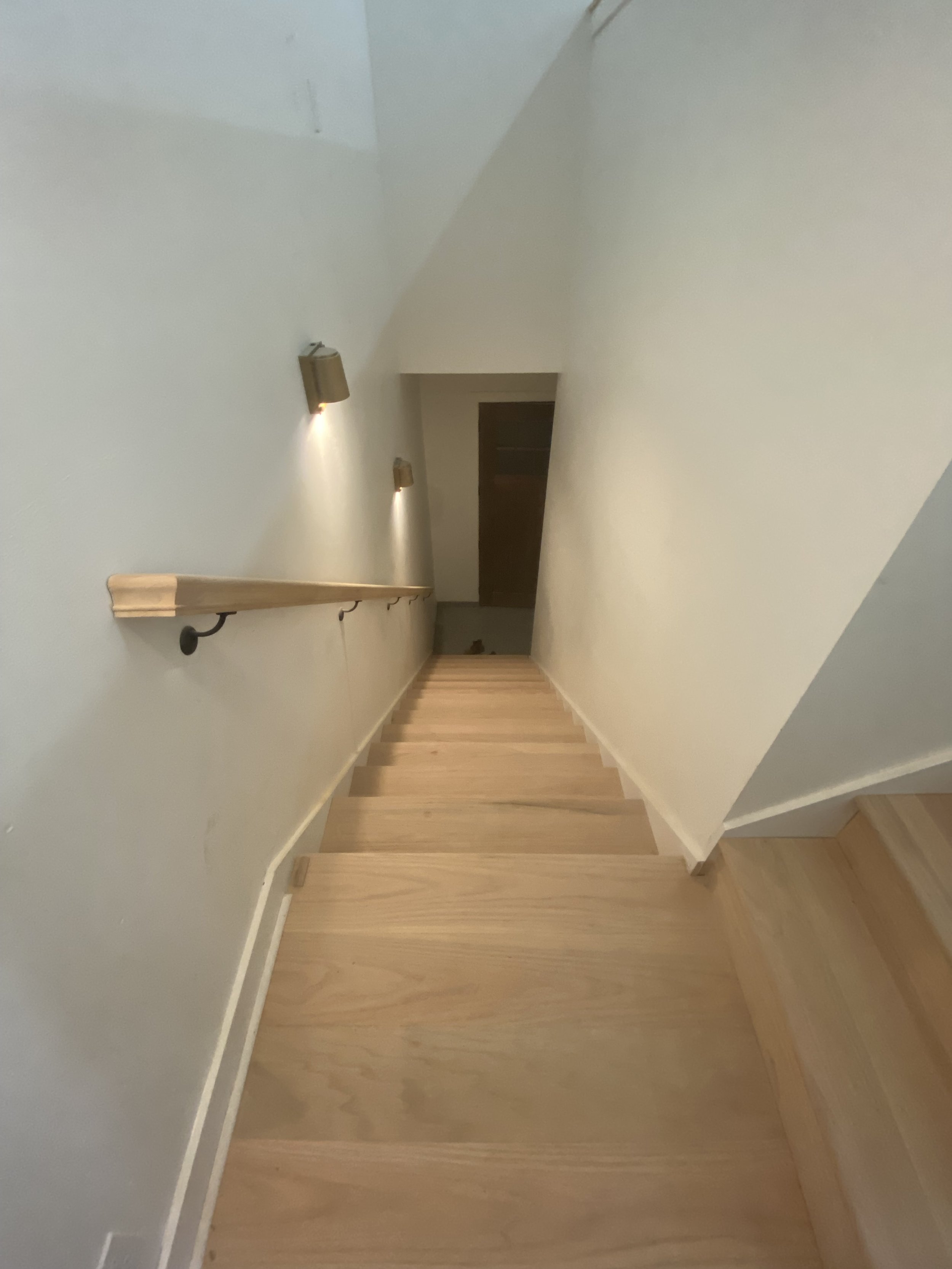 Wooden staircase viewed from above, with a handrail on the left and wall-mounted lights illuminating the stairs, leading down to a door at the bottom.