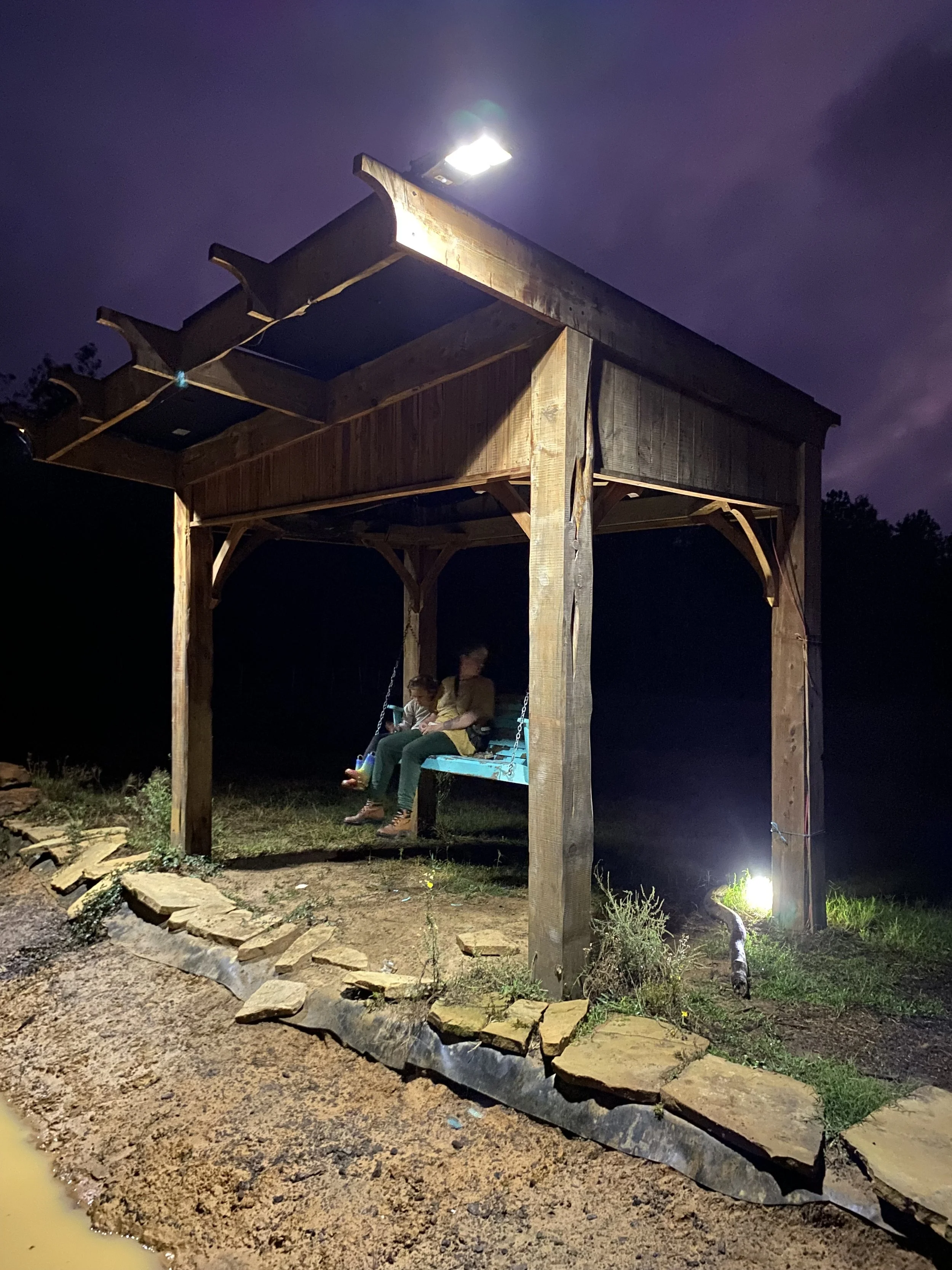 A wooden shelter with a swing bench hanging inside, illuminated by a bright light on a dark evening. Two children are sitting on the swing.