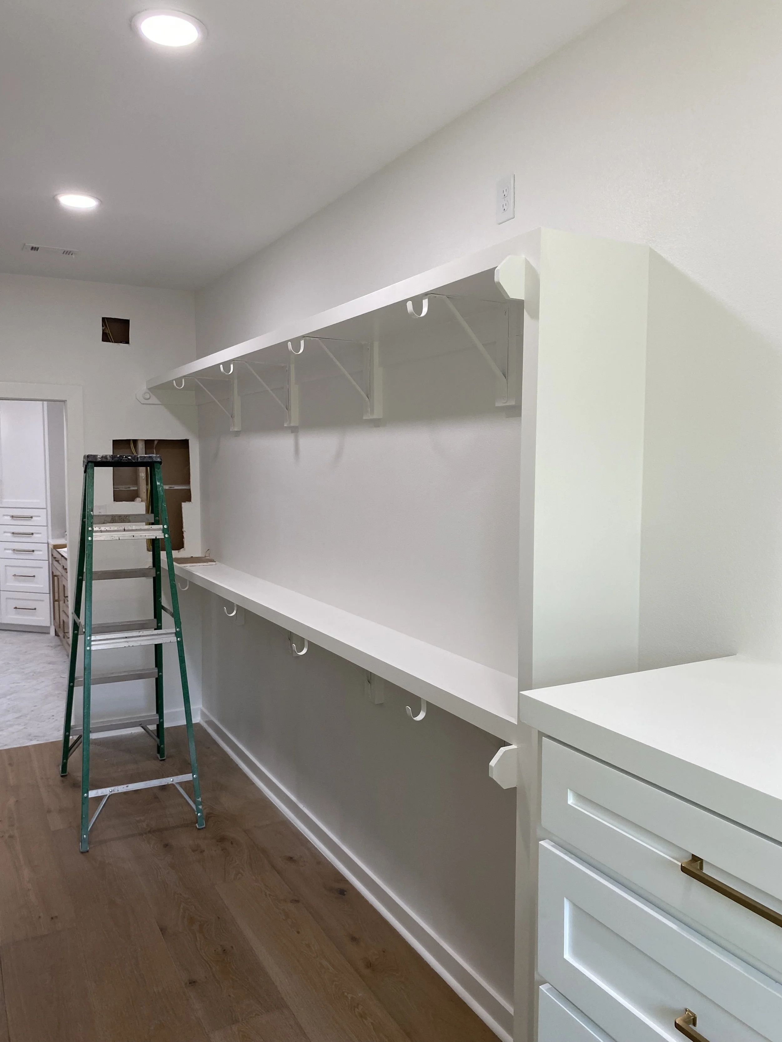 White wall with two-tiered shelves and hooks, a green step ladder, and part of a white cabinet with gold handles on a wooden floor.