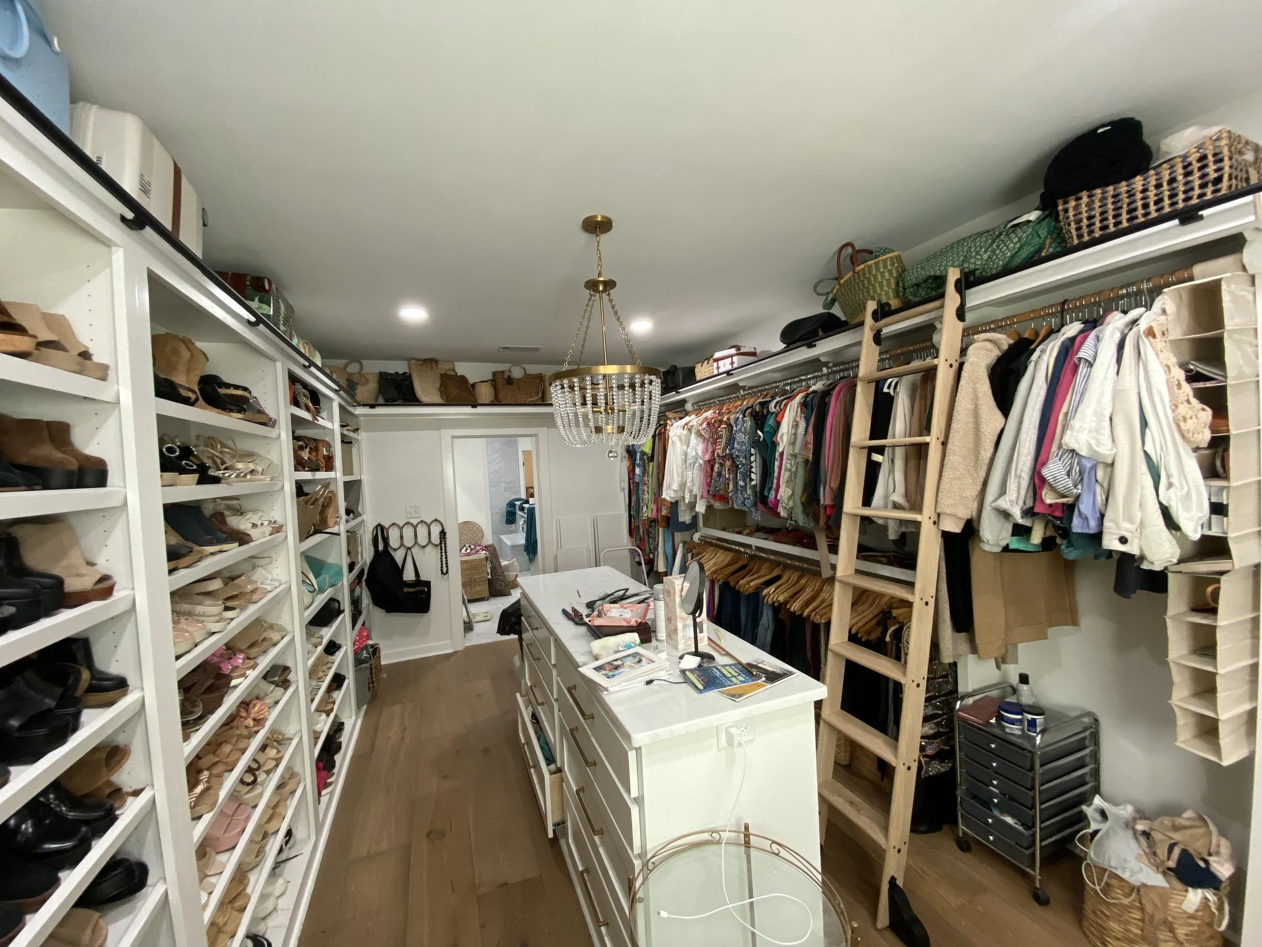 A walk-in closet with shelves of shoes on the left, hanging clothes on the right, a central white dresser with various items on top, and a chandelier hanging from the ceiling.