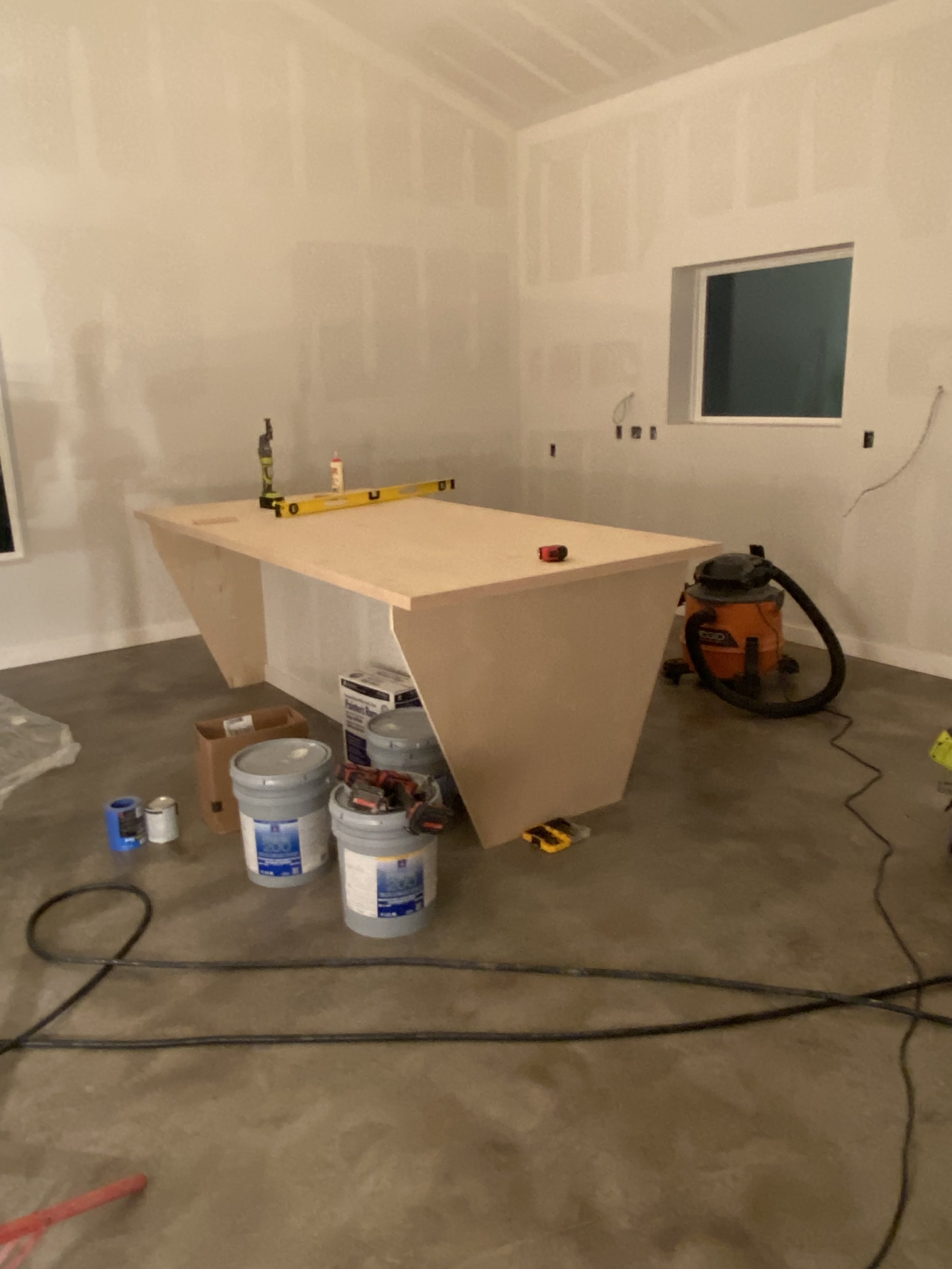 Interior view of a room under construction with a wooden work table, construction tools, buckets, and building supplies scattered on the floor, and a vacuum cleaner against the wall.