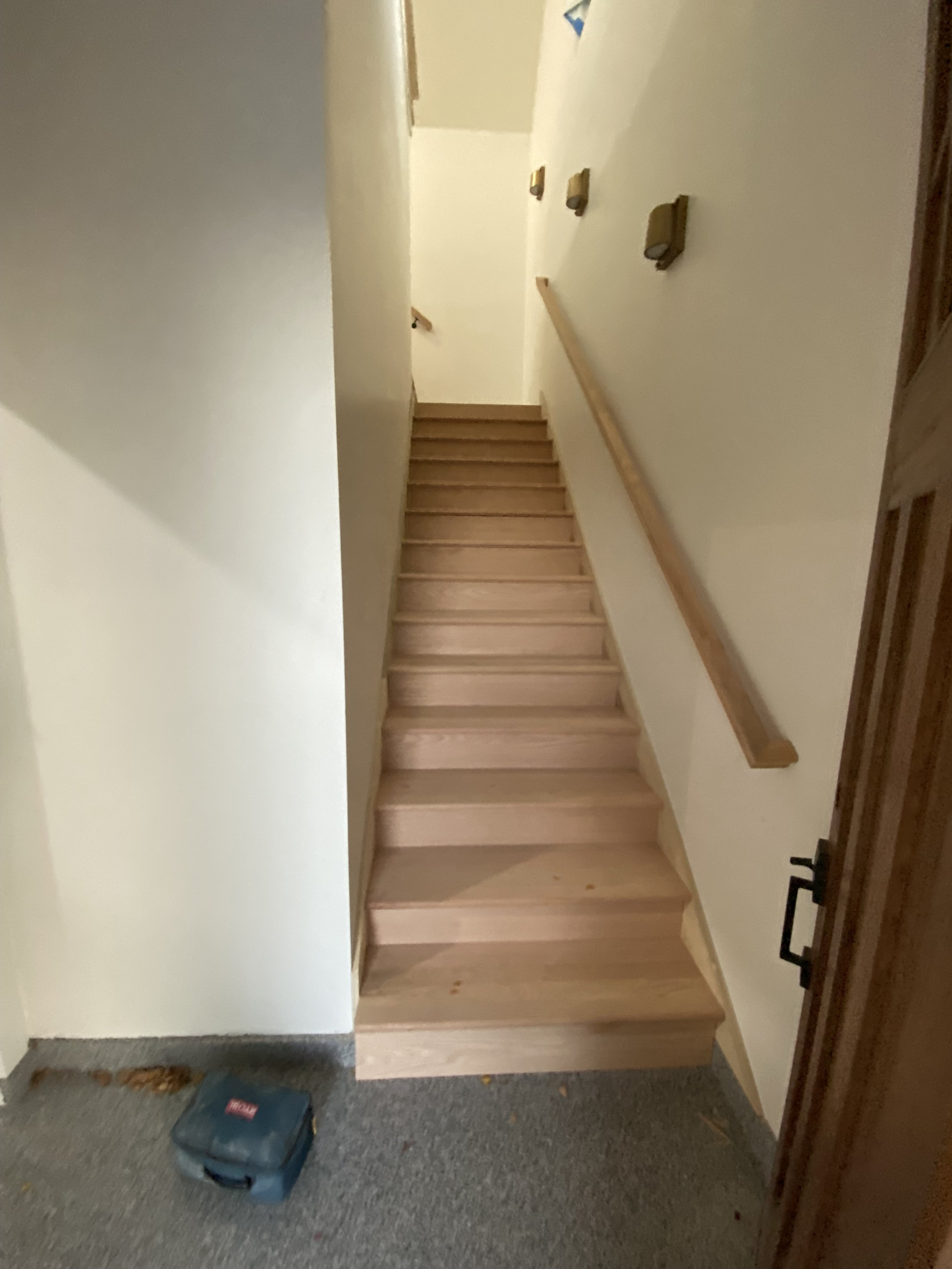 Wooden staircase with a handrail on the right side, leading up to another floor. The stairs are light-colored wood. There are wall-mounted objects on the wall beside the staircase, and a door frame visible to the right. The floor at the bottom of the