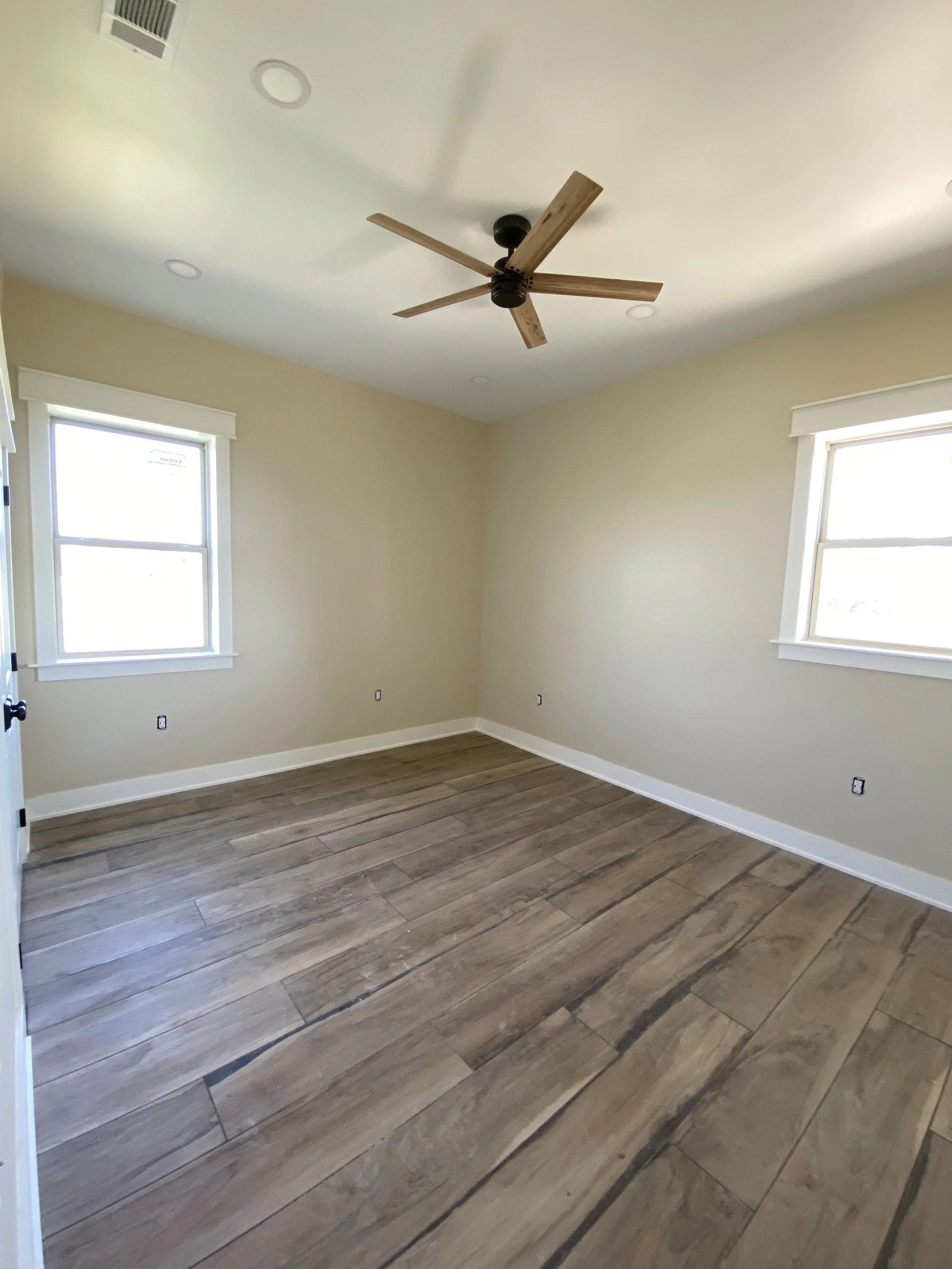 Empty room with two windows, a ceiling fan with wooden blades, beige walls, and wood-look floor tiles.