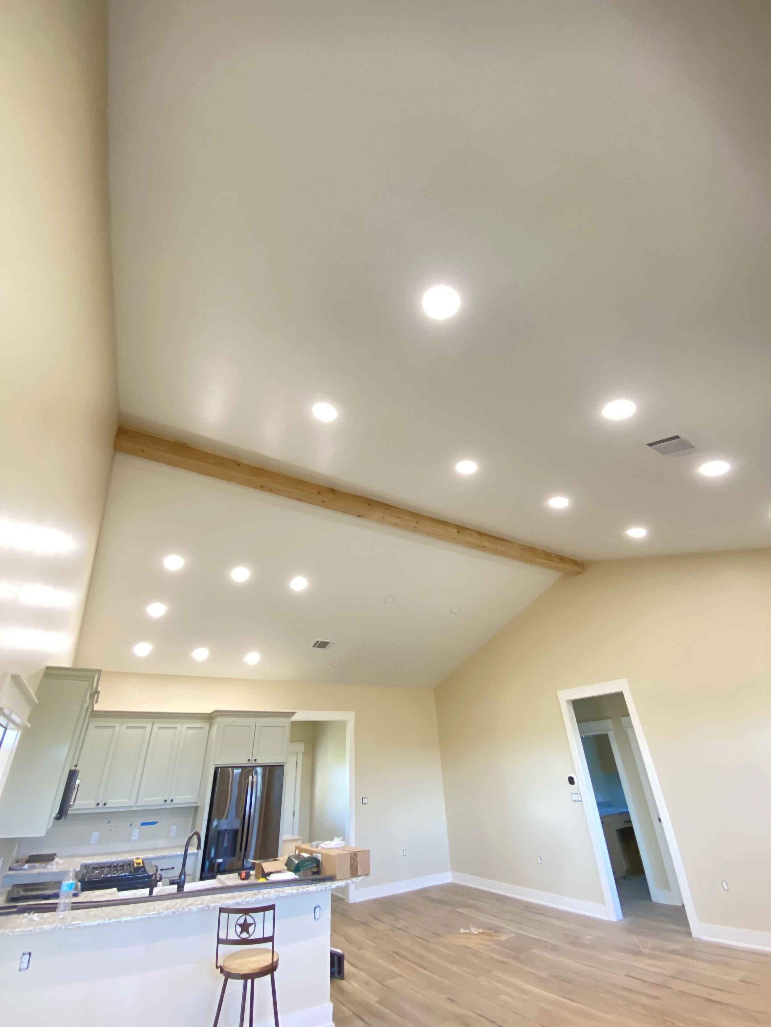 Interior of a living room with a high ceiling, recessed lights, a kitchen with white cabinets, and a wooden beam on the ceiling.