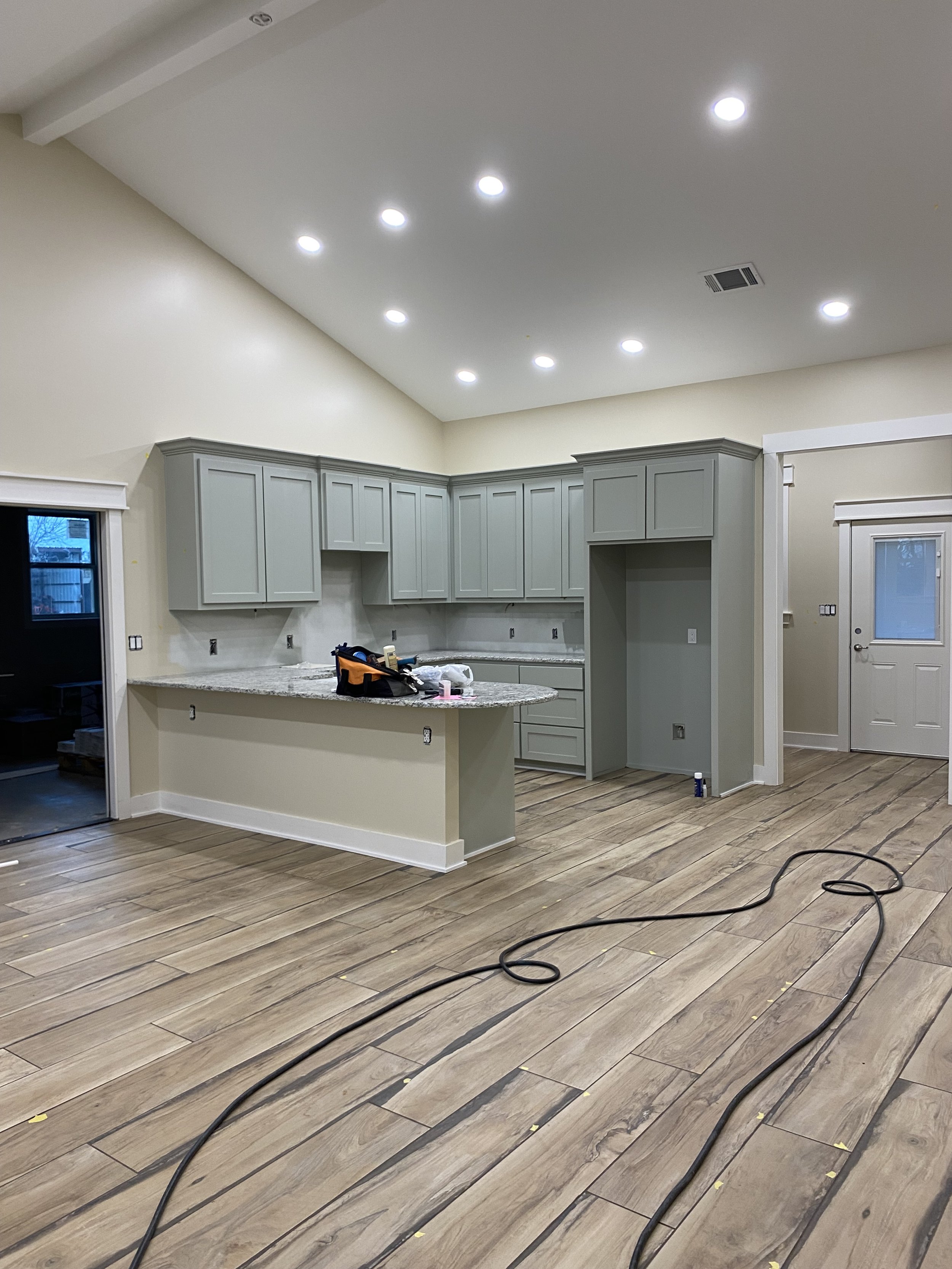 Interior of a kitchen under construction with light wood flooring, gray cabinets, a granite countertop, and ceiling lights, with tools and equipment on the counter.