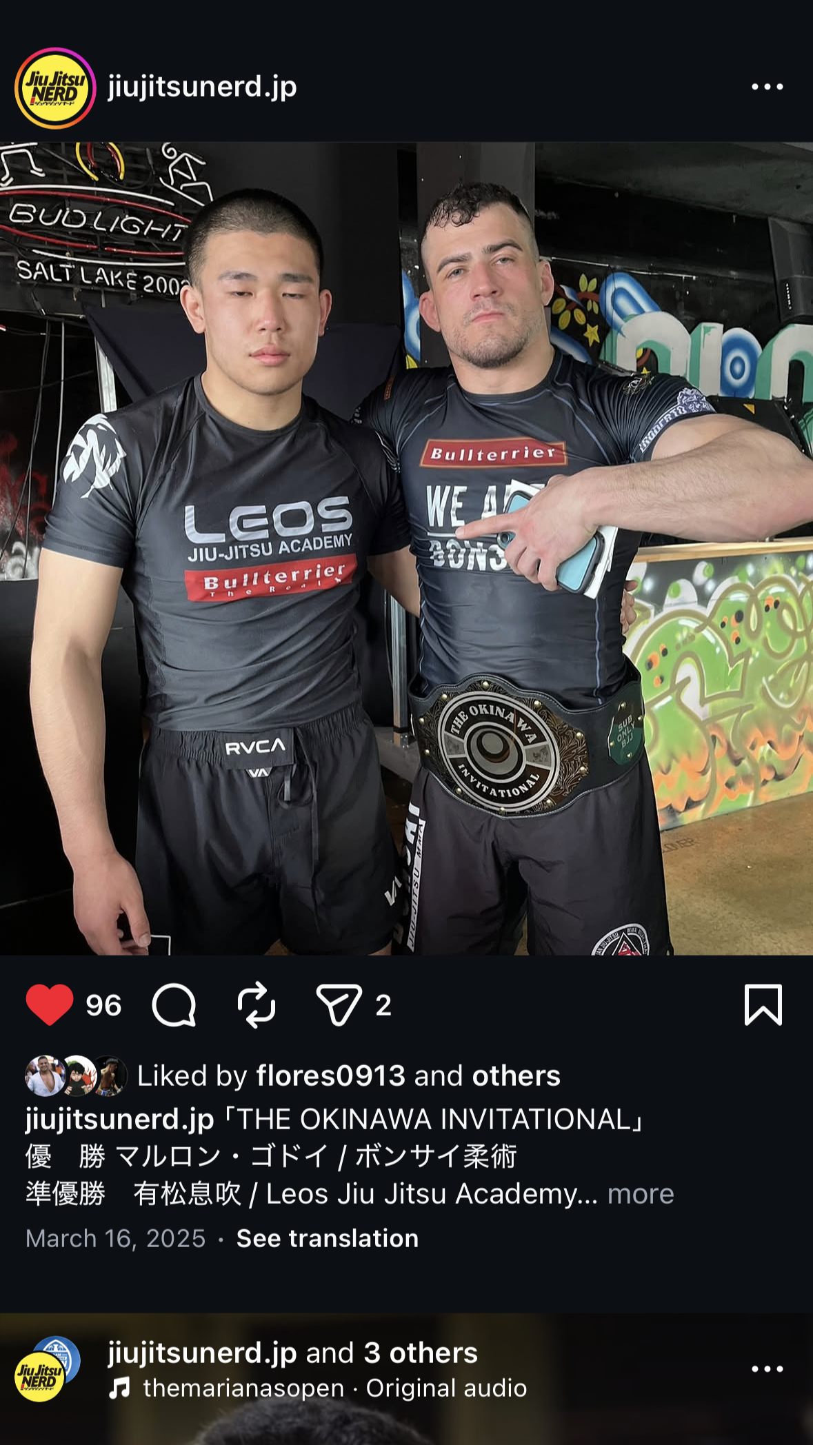 Two men in black athletic shirts and shorts, standing close together. The man on the left has a buzz cut and serious expression. The man on the right has short curly hair, a beard, and is pointing at his championship belt with a stern expression.