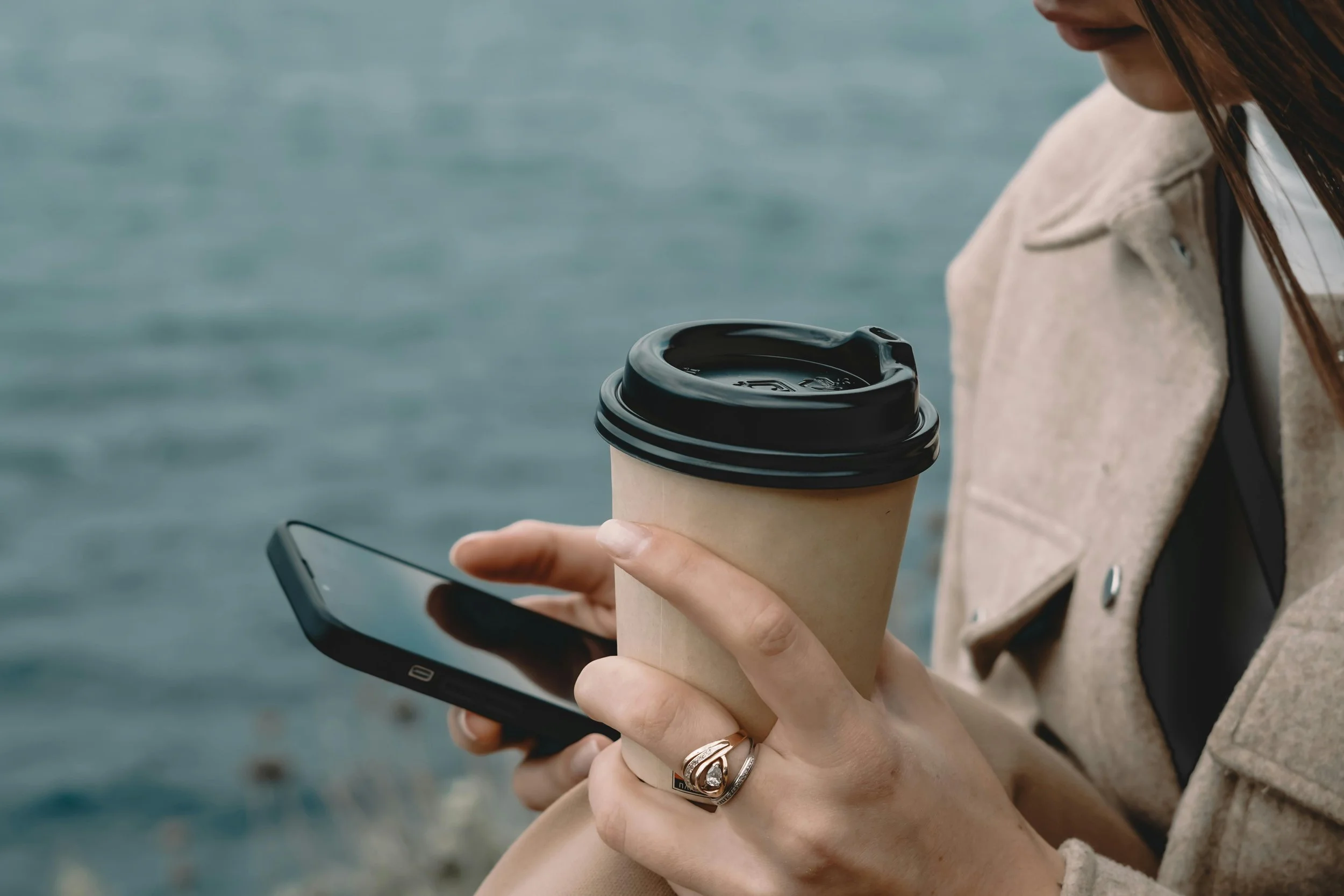 Close-up of a woman's hands holding both a coffee cup and a mobile phone, with the bottom of her mouth and chin visible. She is wearing a tan coat. There appears to be blue calm water in the background.