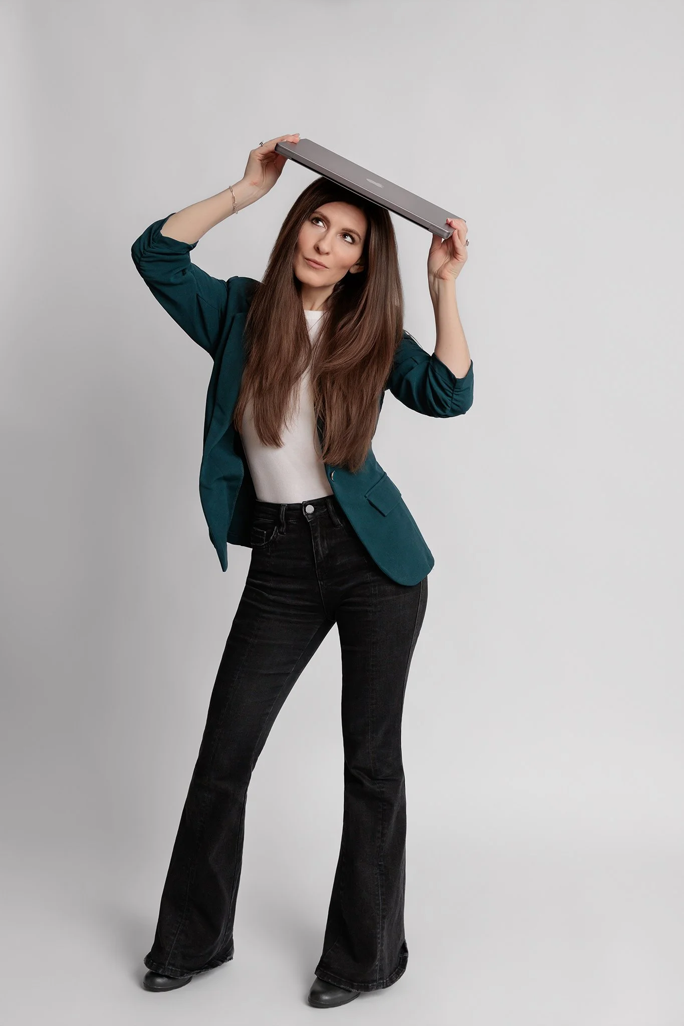 Young woman with long brown hair, wearing black flared jeans, a teal blazer, and white shirt, holding a laptop on top of her head, looking upward and to the side, standing against a plain white background