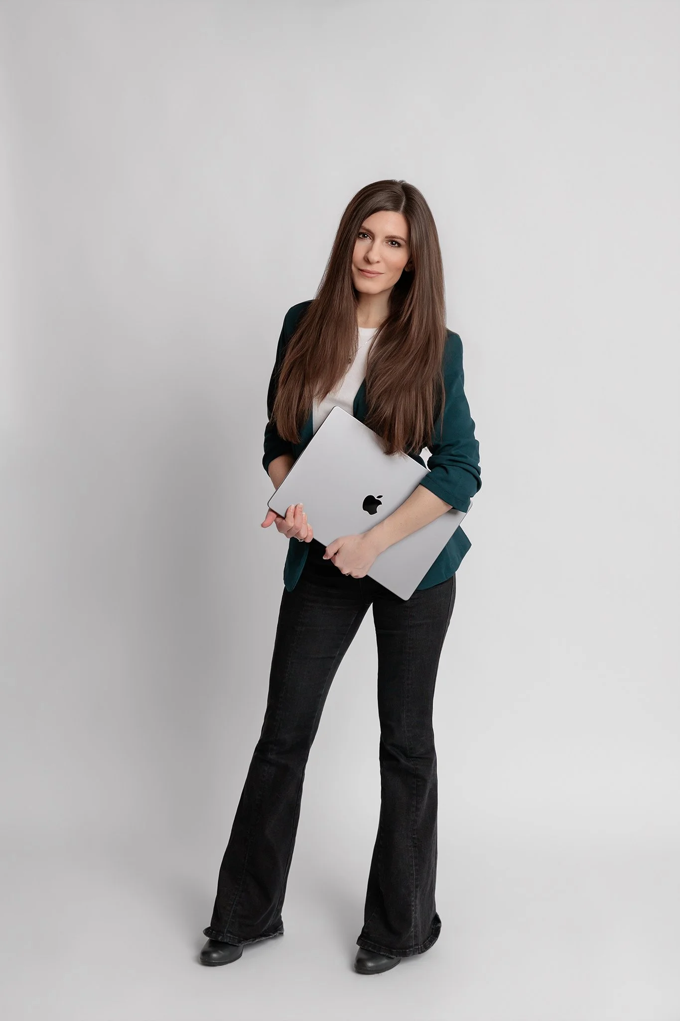 Young woman with long brown hair, wearing a white top and teal blazer and black flared jeans, standing against a plain white background and holding a Macbook laptop