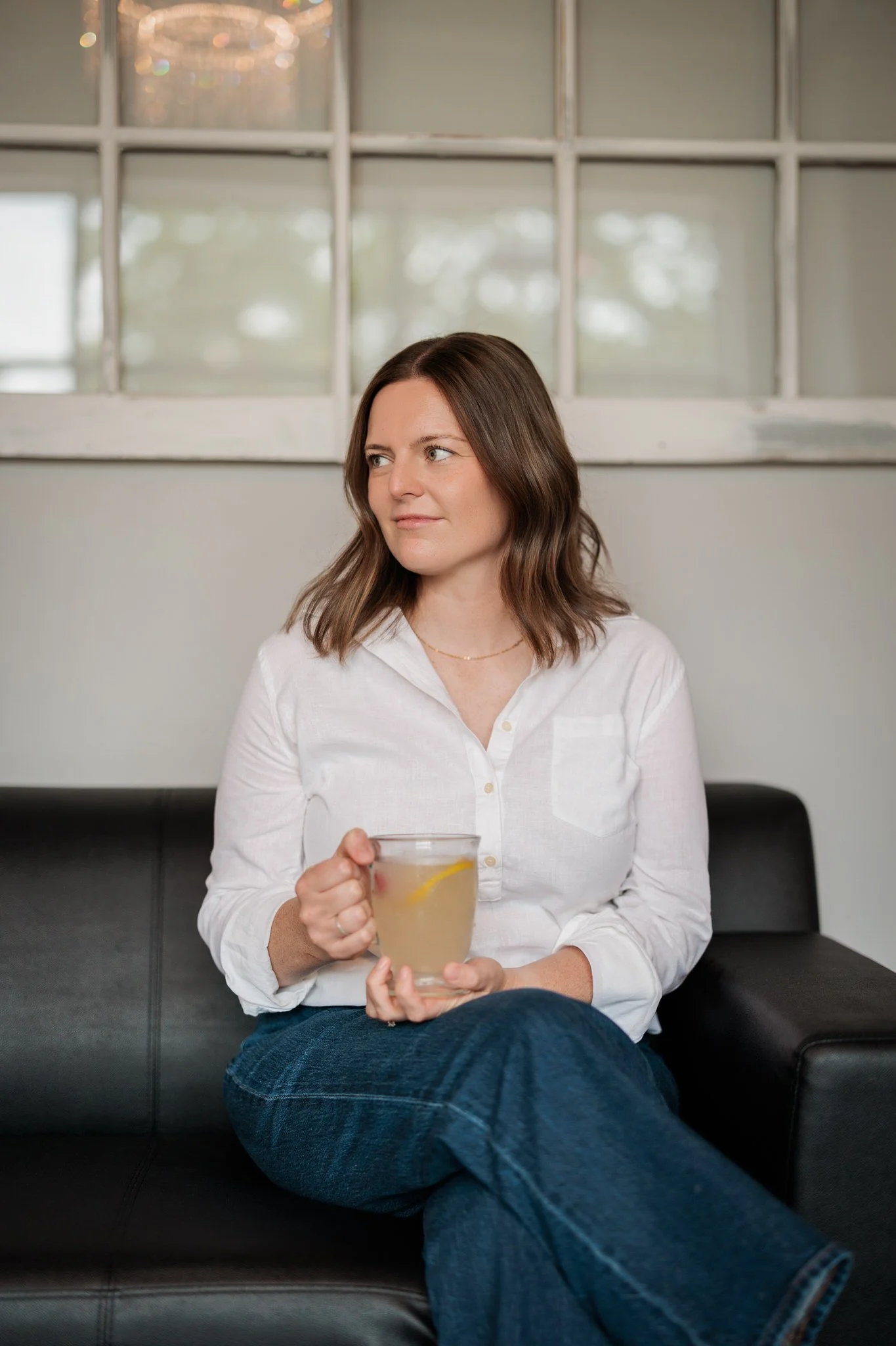 Woman with brown hair and a white shirt and jeans, sitting on a black couch, holding a glass of lemonade and looking to the side