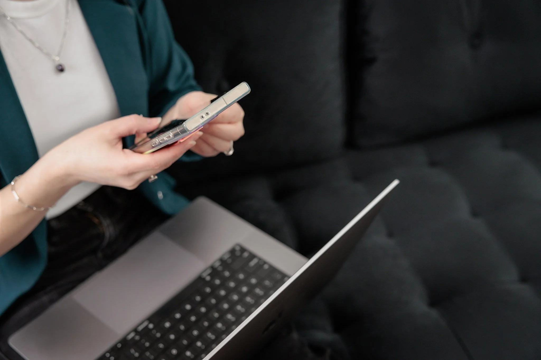 Close-up of a woman wearing a white top and teal green blazer, holding a mobile phone, with a laptop open on her lap. She is sitting on a black tufted couch.