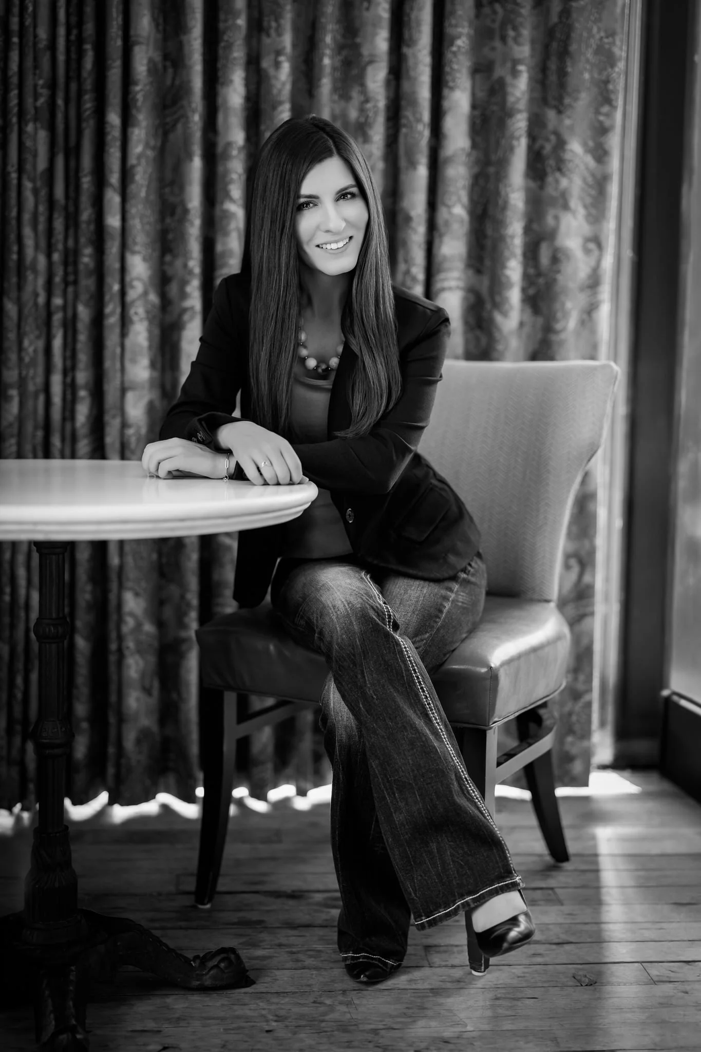 Roxanne Abler sitting at a round cafe table, wearing a black blazer and jeans, image in black and white