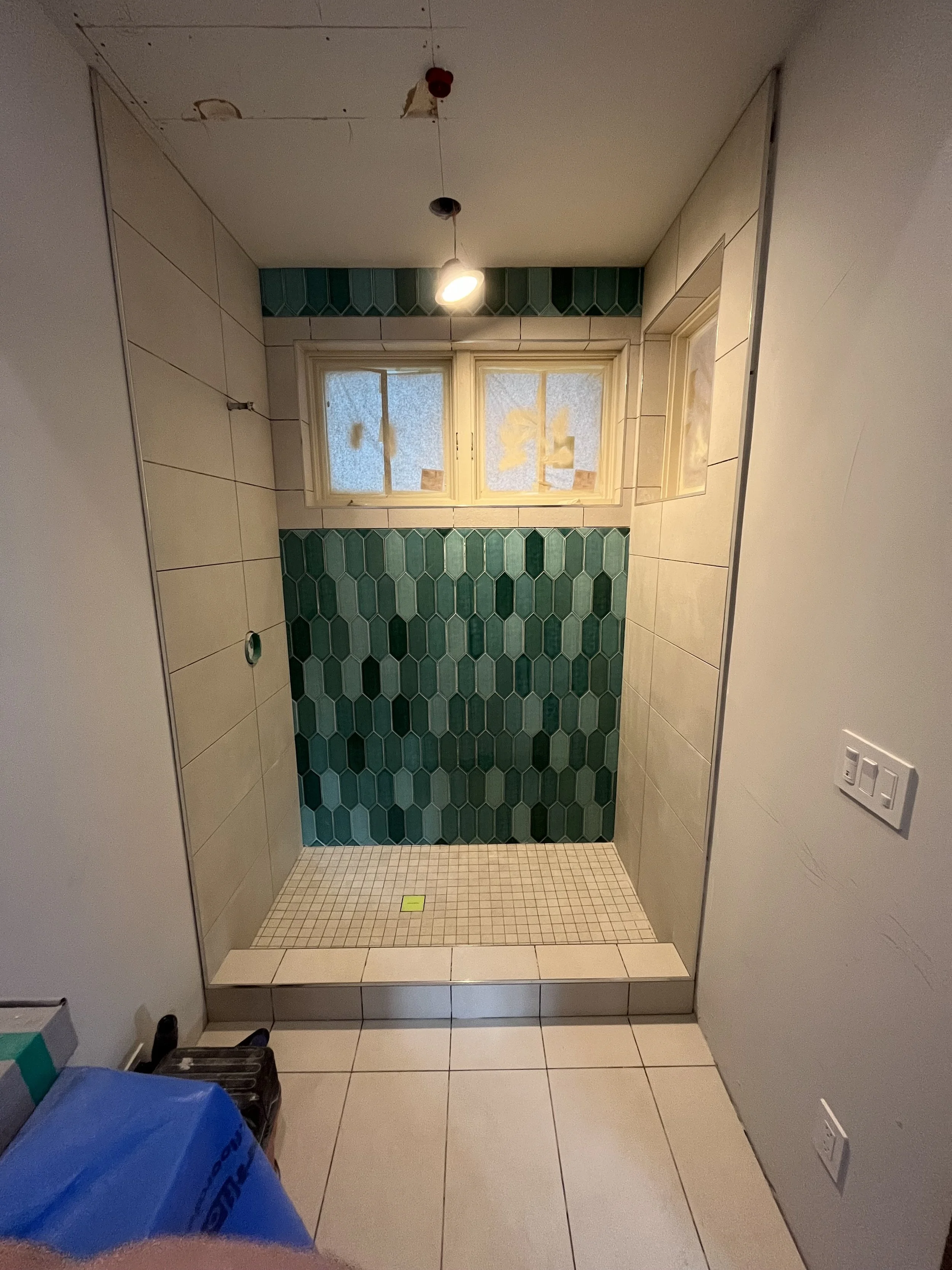 Bathroom shower area under construction with green hexagonal tiles on the back wall, beige tiles on the side walls, unfinished ceiling, and a small window at the top.