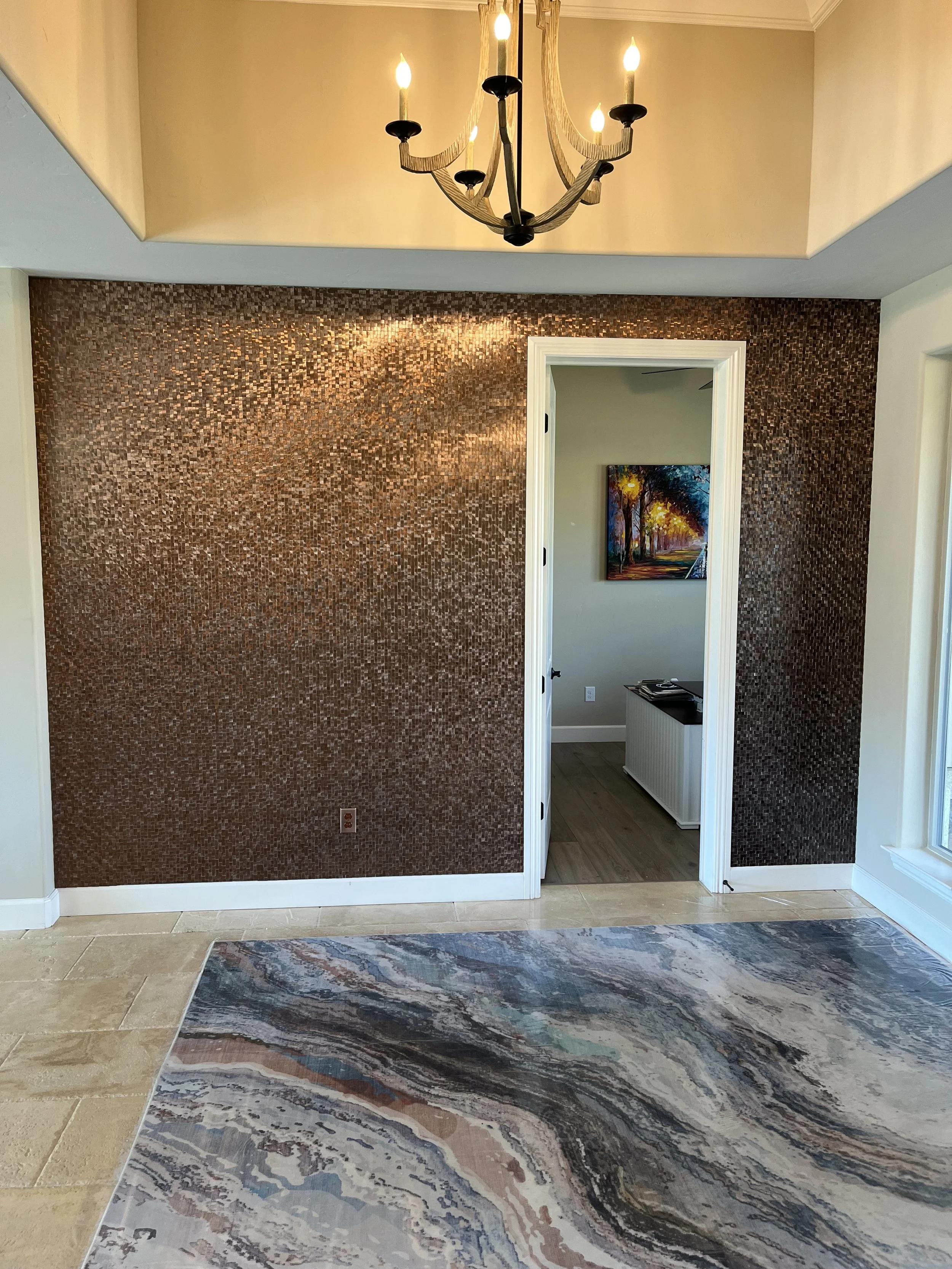 An interior room with a copper mosaic accent wall, a chandelier with five lit bulbs hanging from a high ceiling, a colorful landscape painting on the wall in the adjacent room, a large window on the right, and a marble-patterned rug on light-colored tiled flooring.