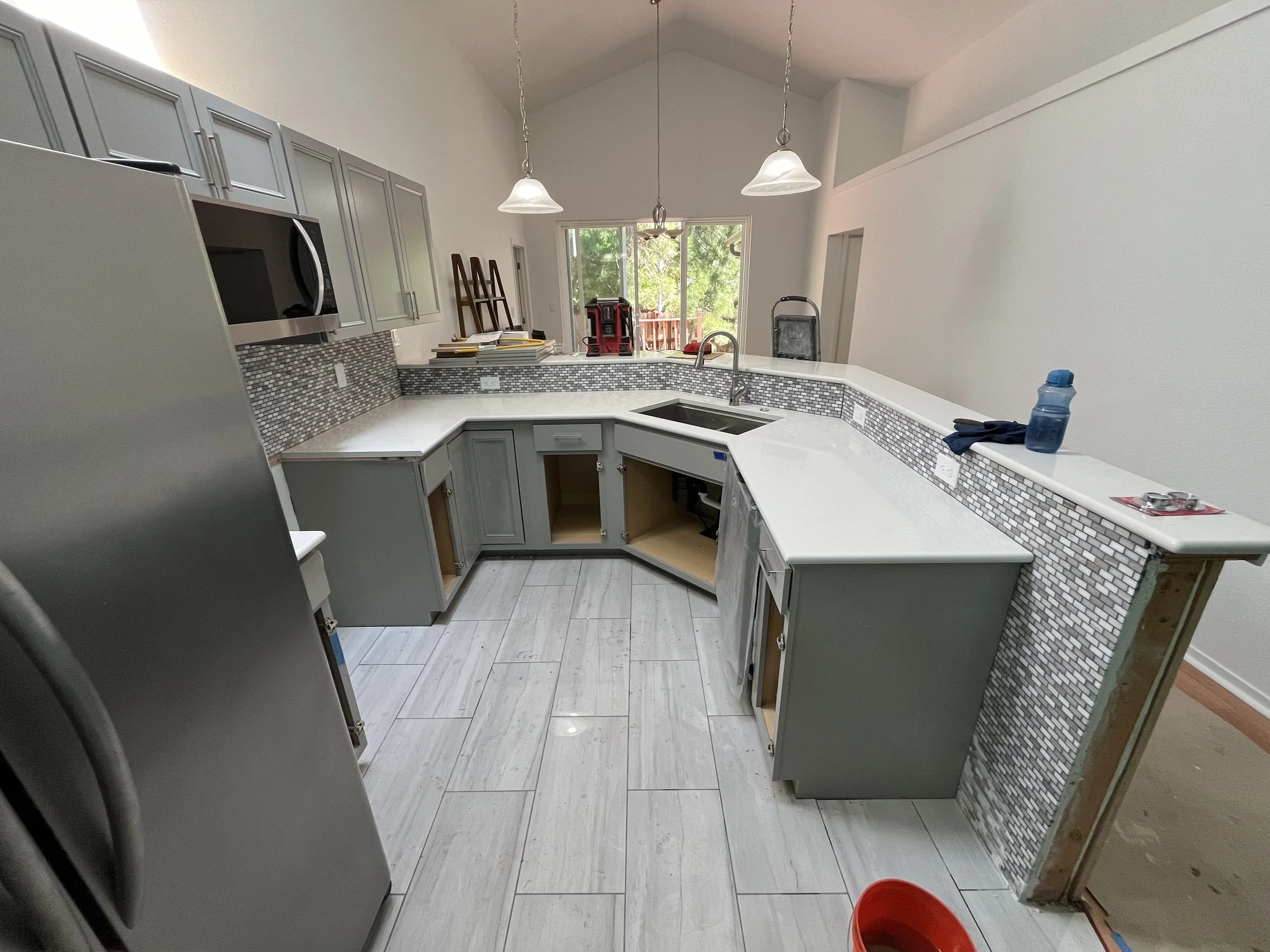 Kitchen under construction with gray cabinets, mosaic tile backsplash, white countertops, a stainless steel microwave, and a window overlooking a backyard.