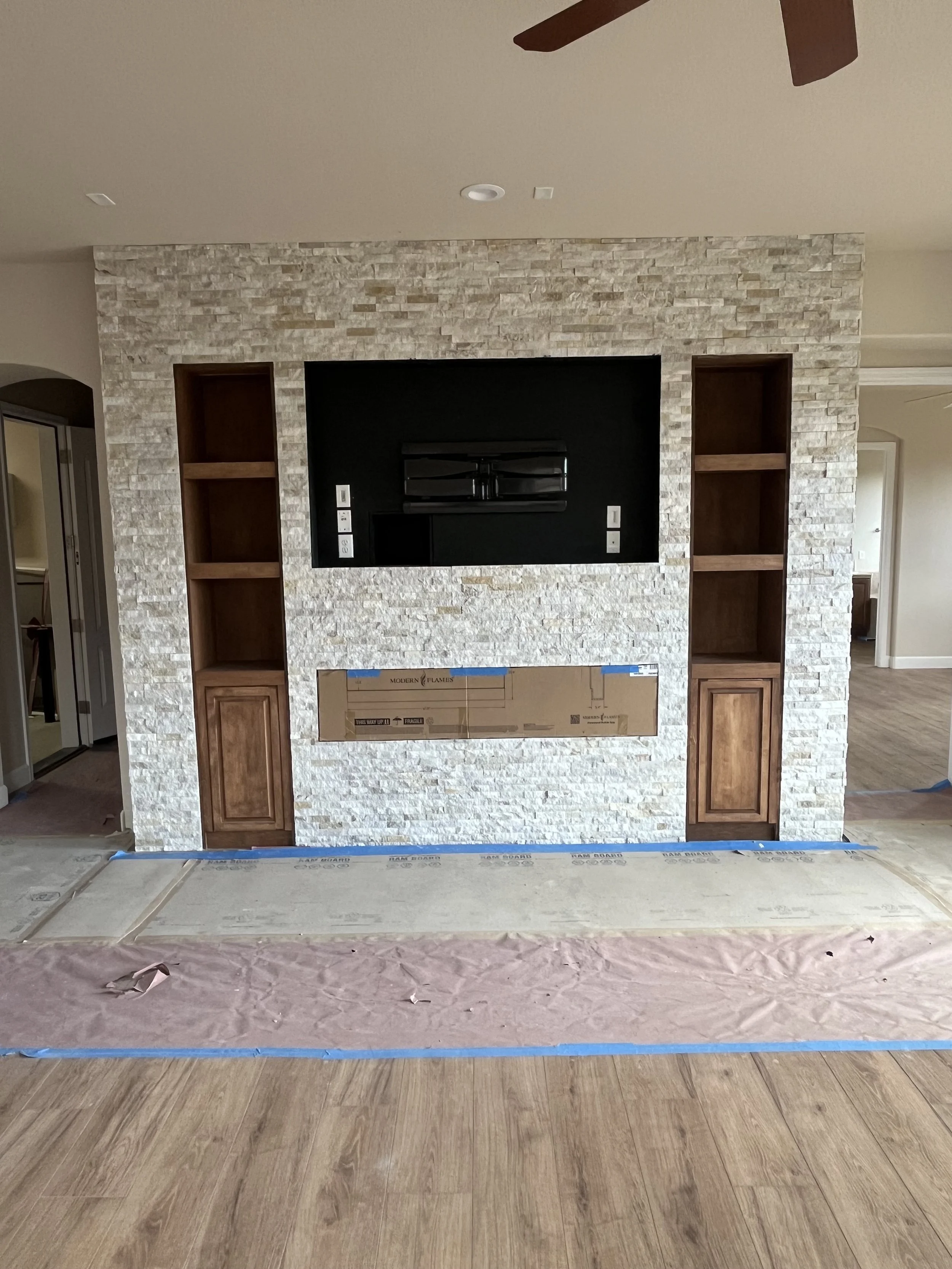 Living room fireplace with a stone surround, built-in wooden shelves on each side, and a black TV mount or insert in the center. The floor has a protective covering, indicating ongoing renovation or installation work.