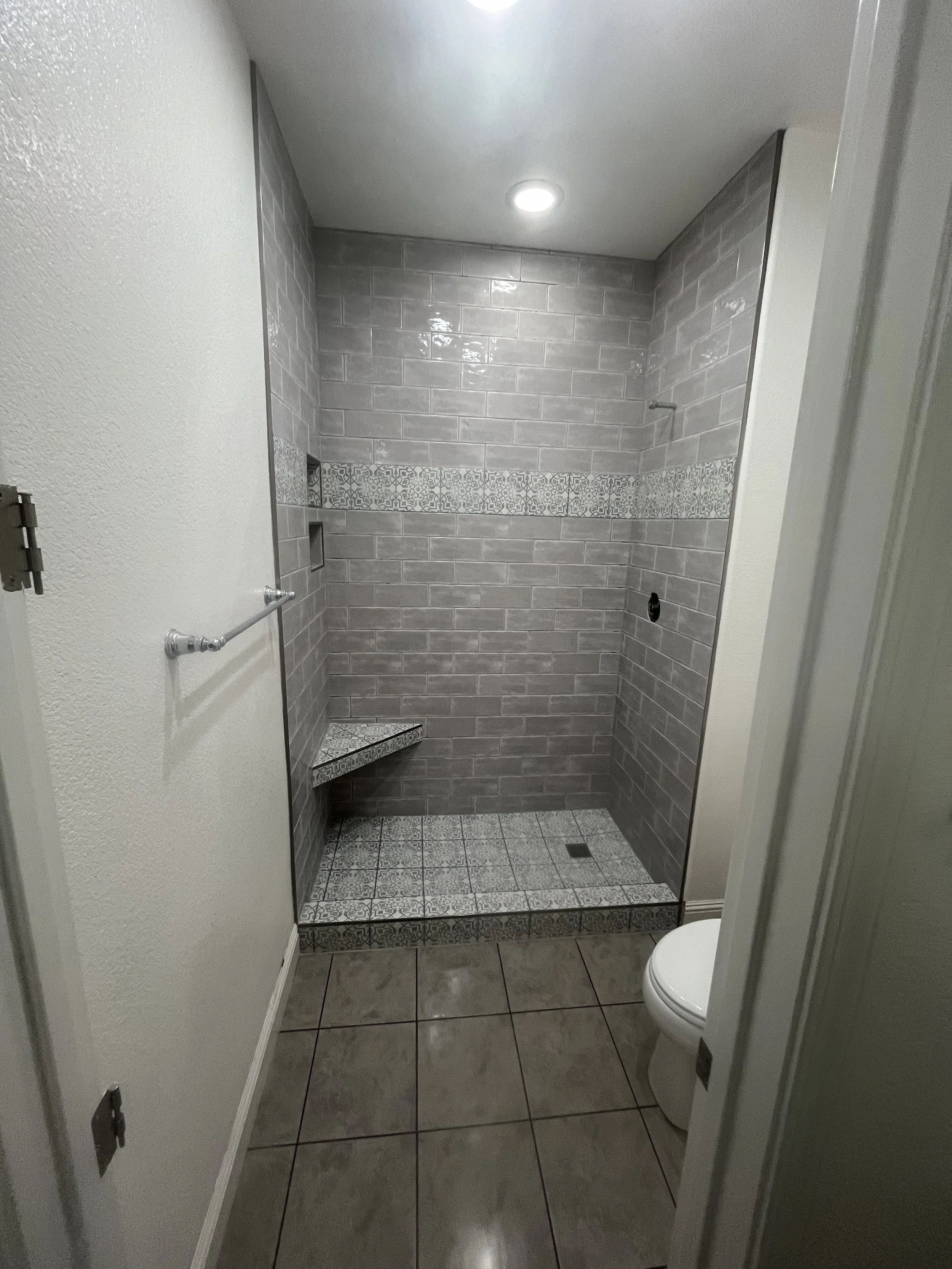 Empty walk-in shower with gray tiled walls, decorative tile strip, built-in corner bench, and gray tiled floor in a bathroom.