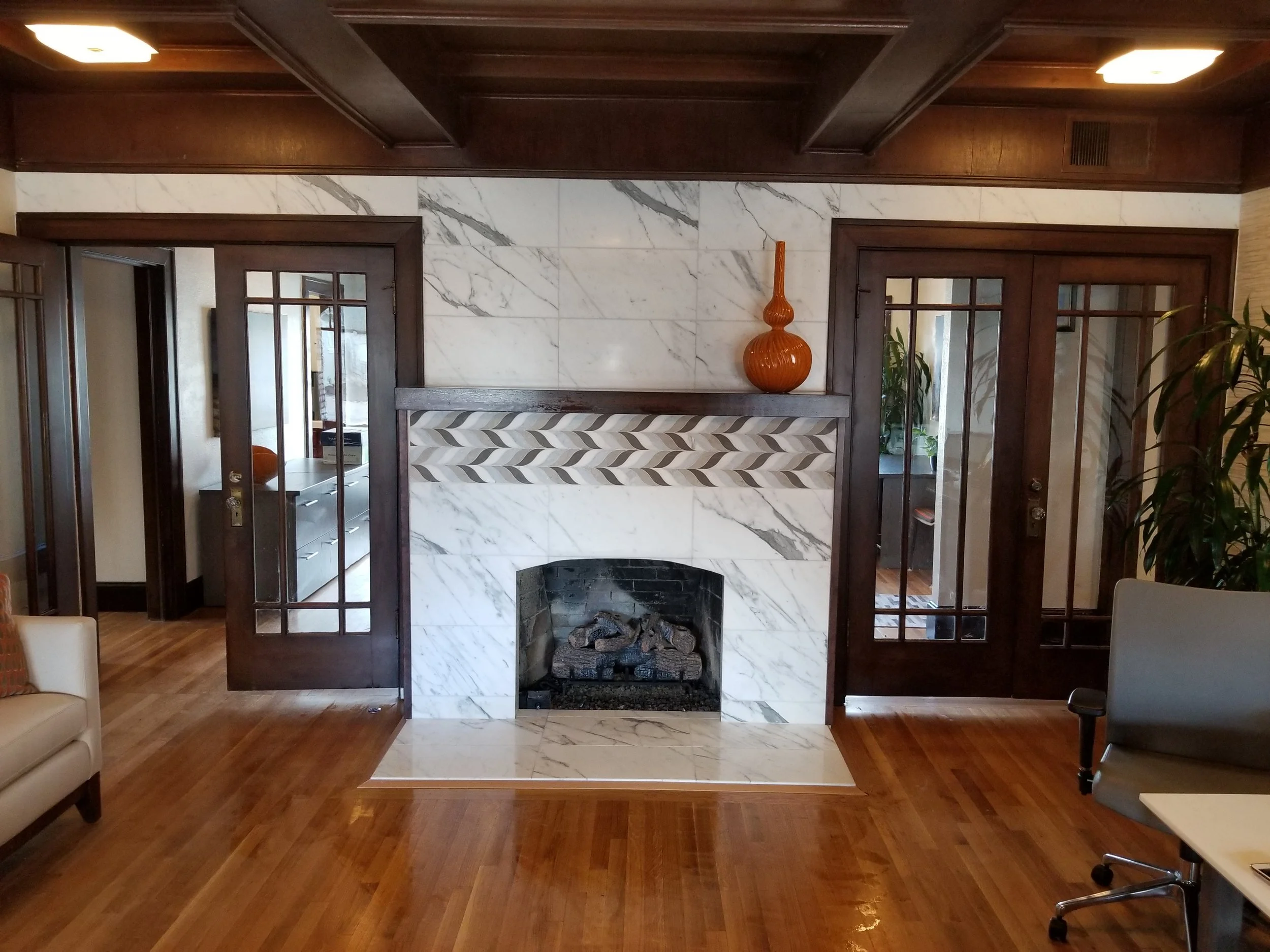 Living room with a white marble fireplace, dark wood walls, and hardwood floors. Two glass-paneled wooden doors on either side of the fireplace. An orange decorative vase on the mantel and a large indoor plant on the right.
