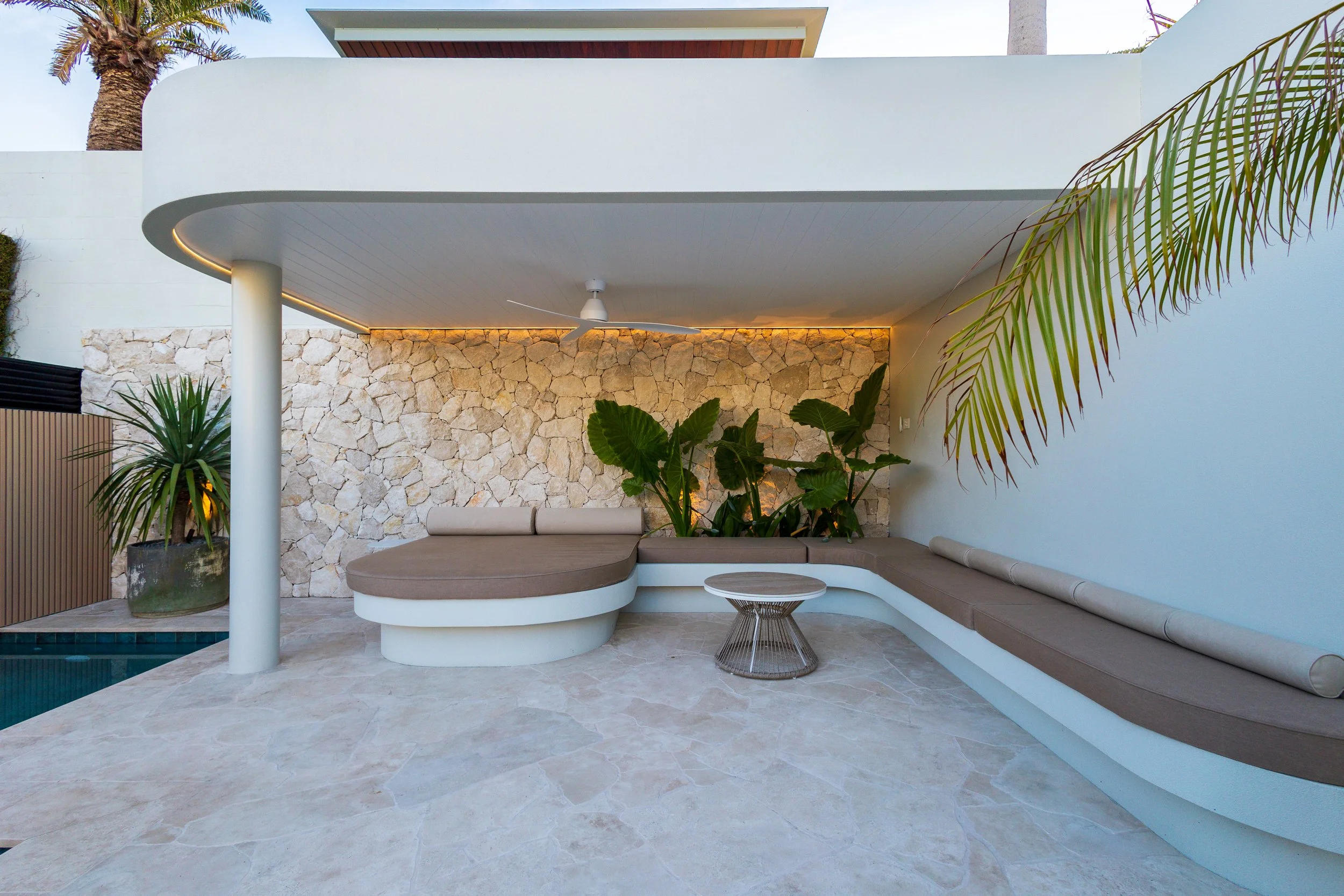 Modern outdoor seating area with crazy pave feature wall and french pattern pool surrounds, curved built-in sofa, a small round coffee table, potted plants, a stone wall, and a ceiling fan with a white ceiling and wall, surrounded by palm trees.