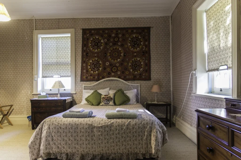 A cozy bedroom with floral wallpaper, a bed with green and floral pillows, a tapestry above the bed, two windows with roller shades, a small nightstand with a lamp, a dresser, and a laundry basket.