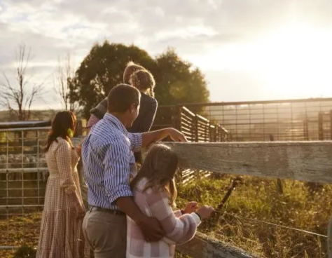 A group of people, including children and adults, at a farm standing by a wooden fence during sunset, observing or interacting with animals.