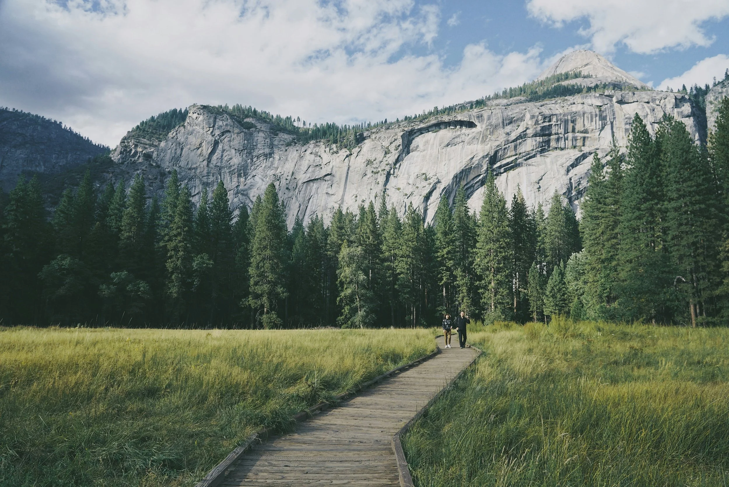 Two people walk on a wooden trail through tall green grass in a forested valley with towering granite cliffs and pine trees in the background, under a partly cloudy sky.