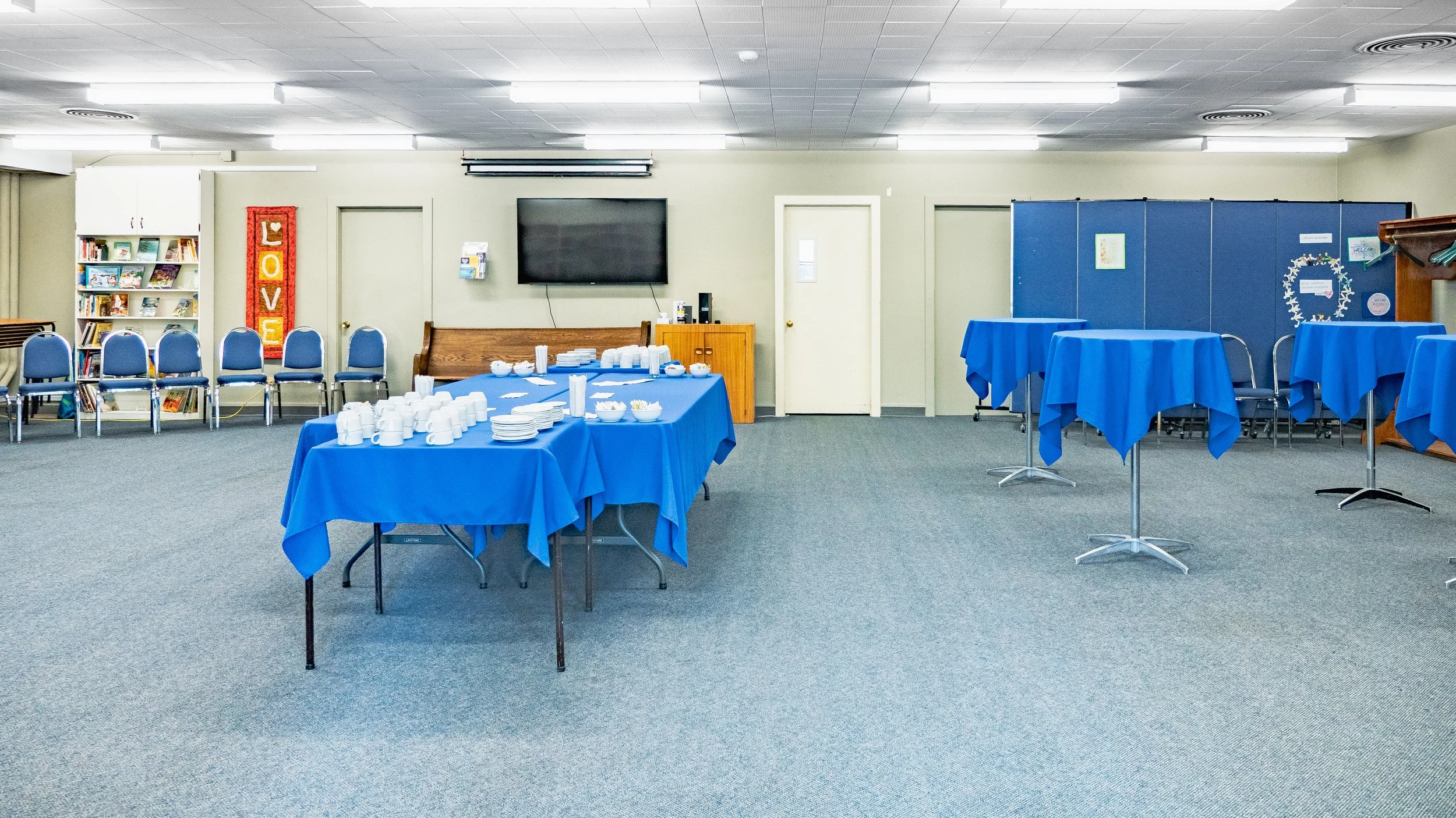 Room set up with round tables covered with blue tablecloths, cups and plates, chairs against the wall, and a refreshment area with cups and snacks. Bookshelf, TV, and decorative hall on the wall.