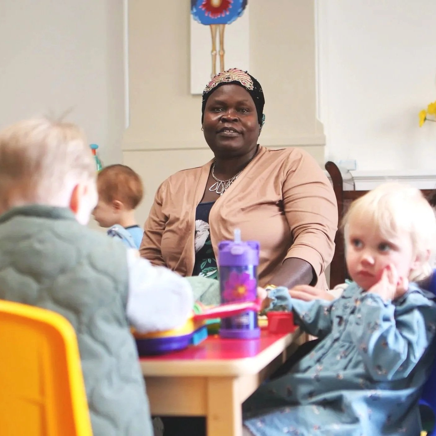 A woman with dark skin, wearing a light brown jacket and a floral headband, sits at a table with young children in a nursery setting. The children are engaged in activities at the table, with one girl looking at the camera.