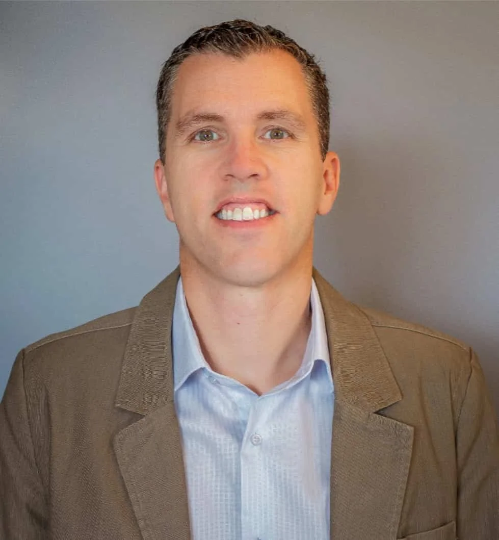 A man with short dark hair, light skin, wearing a brown blazer and a light-colored collared shirt, standing against a grey background.