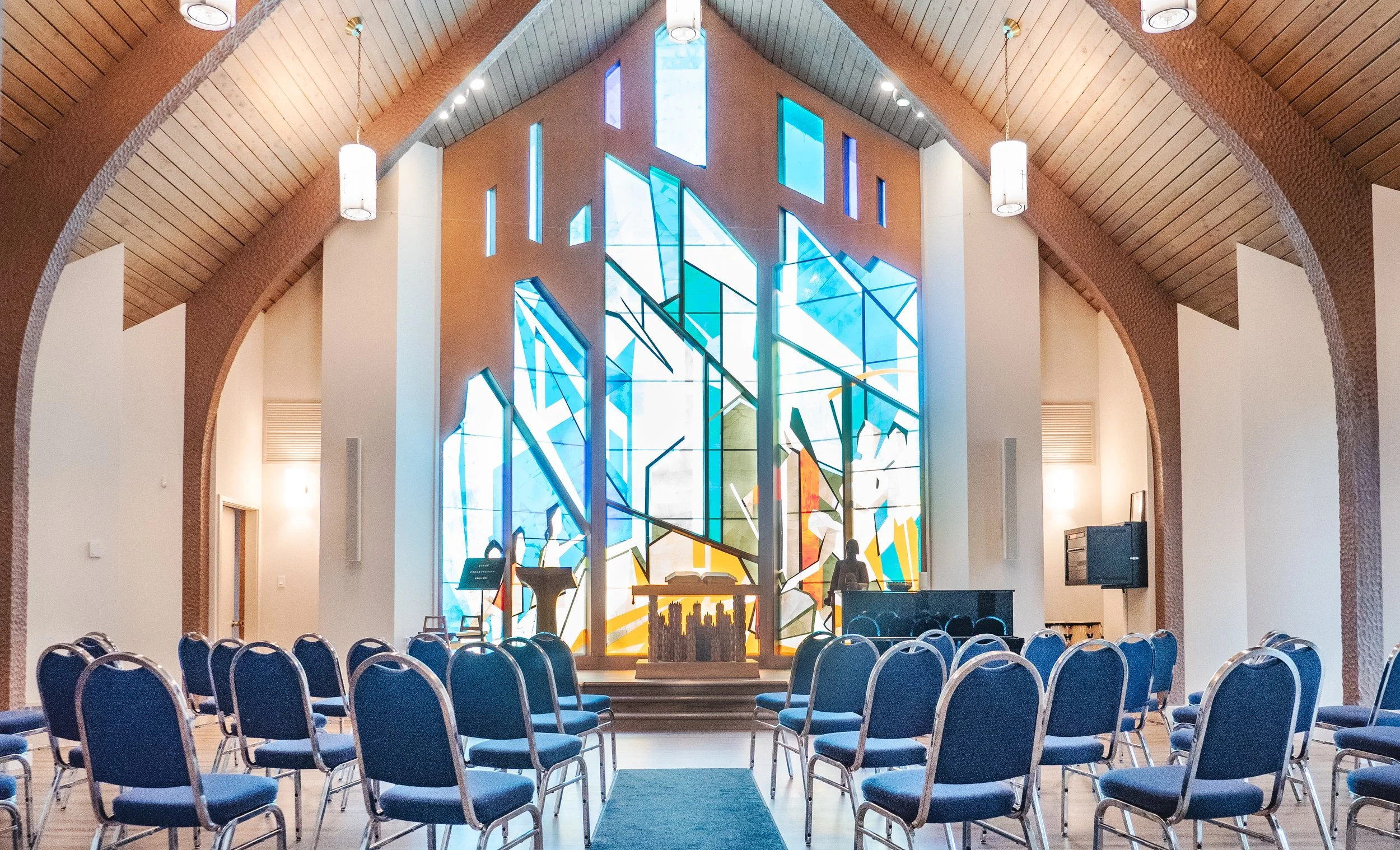Interior of a church with blue chairs arranged in rows facing a modern stained glass window featuring abstract blue, orange, yellow, and green shapes, with an altar and two statues in front.