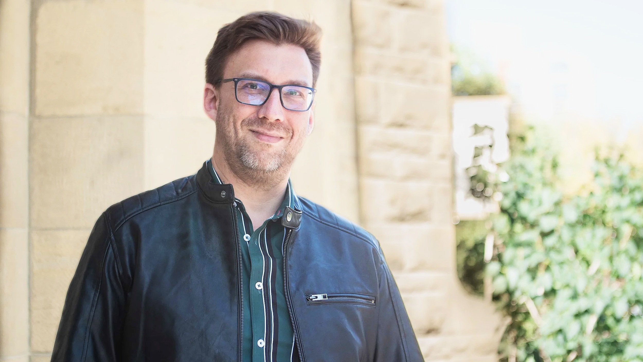 Smiling man with glasses wearing a black leather jacket standing outdoors near a stone wall with greenery in the background.