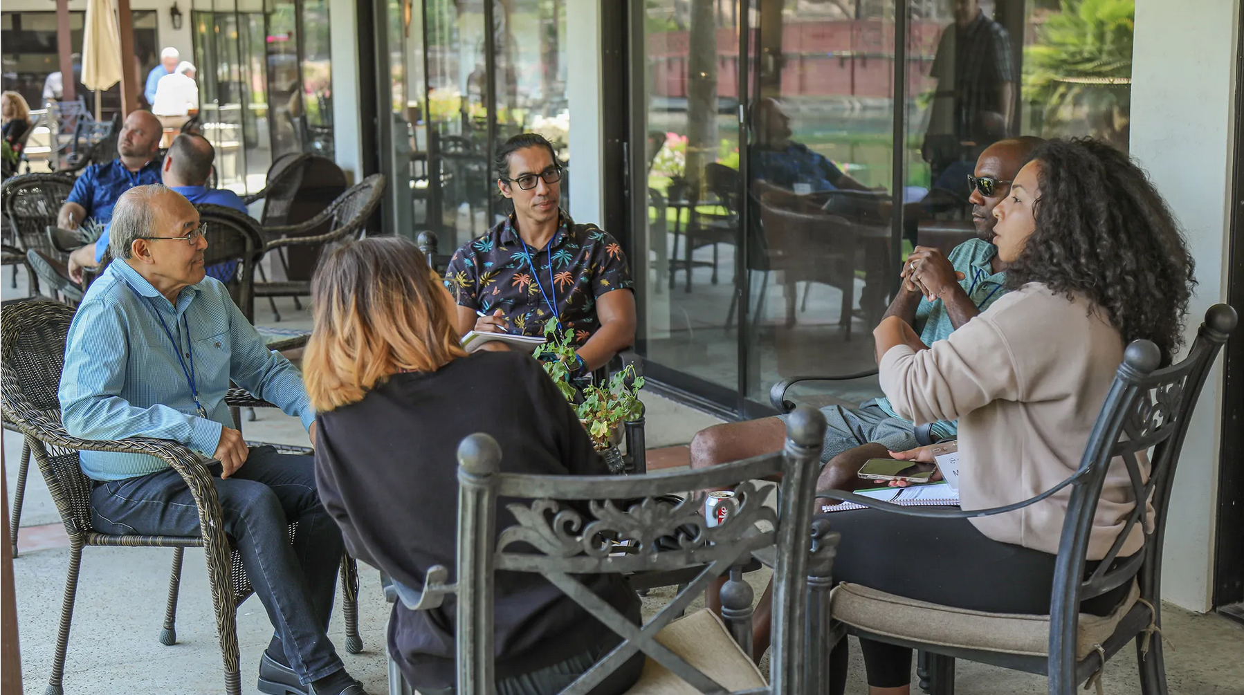 Group of six people sitting in a circle on outdoor patio, engaged in discussion, with coffee cups, notebooks, and a plant on the table in front of them.