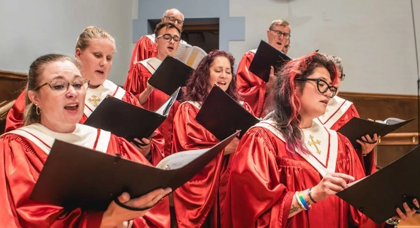 A choir of people singing in a church, wearing red robes with white collars and holding black sheet music.