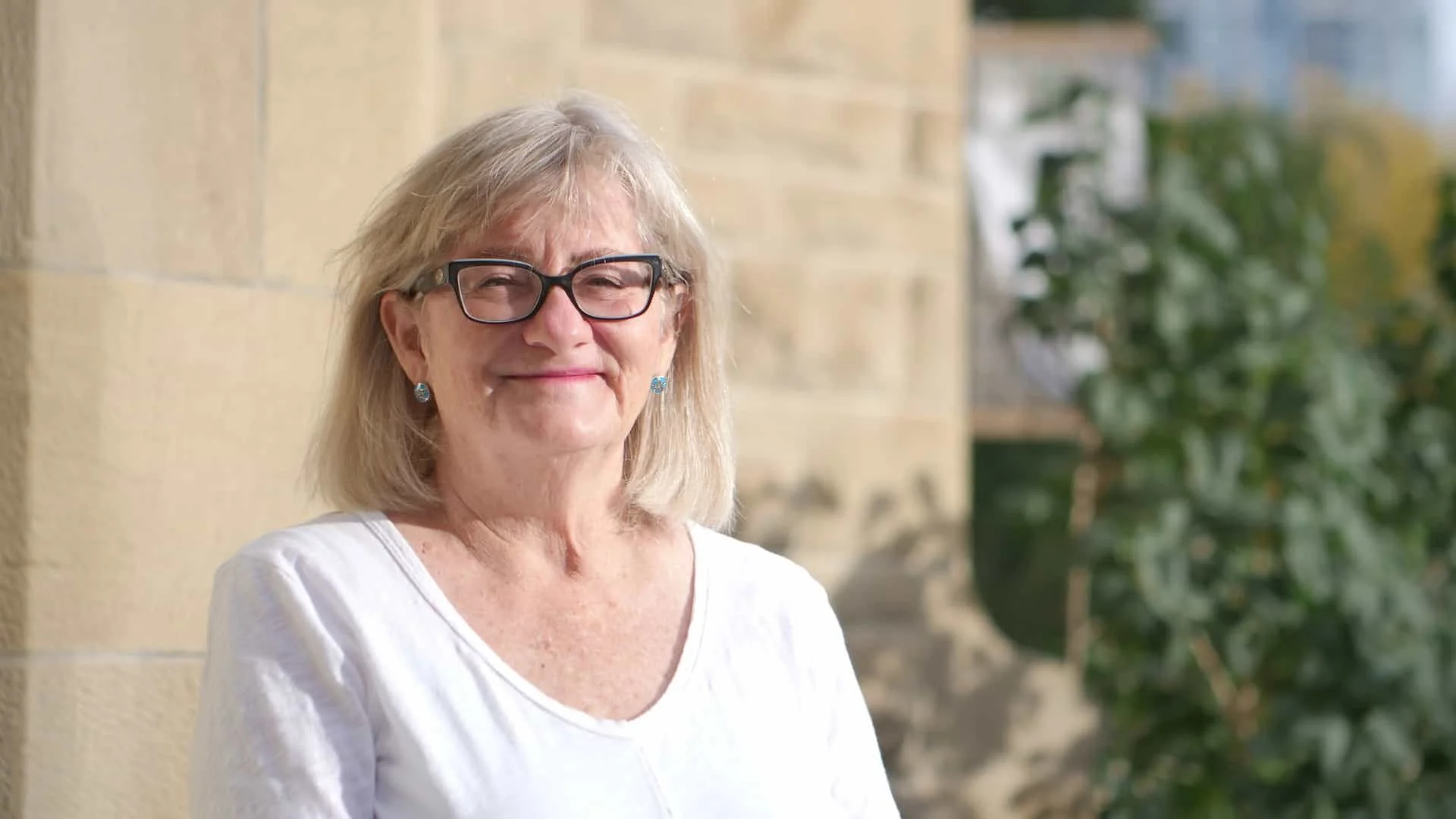 Smiling elderly woman with glasses and earrings, wearing a white shirt, standing outdoors near a brick wall with greenery in the background.