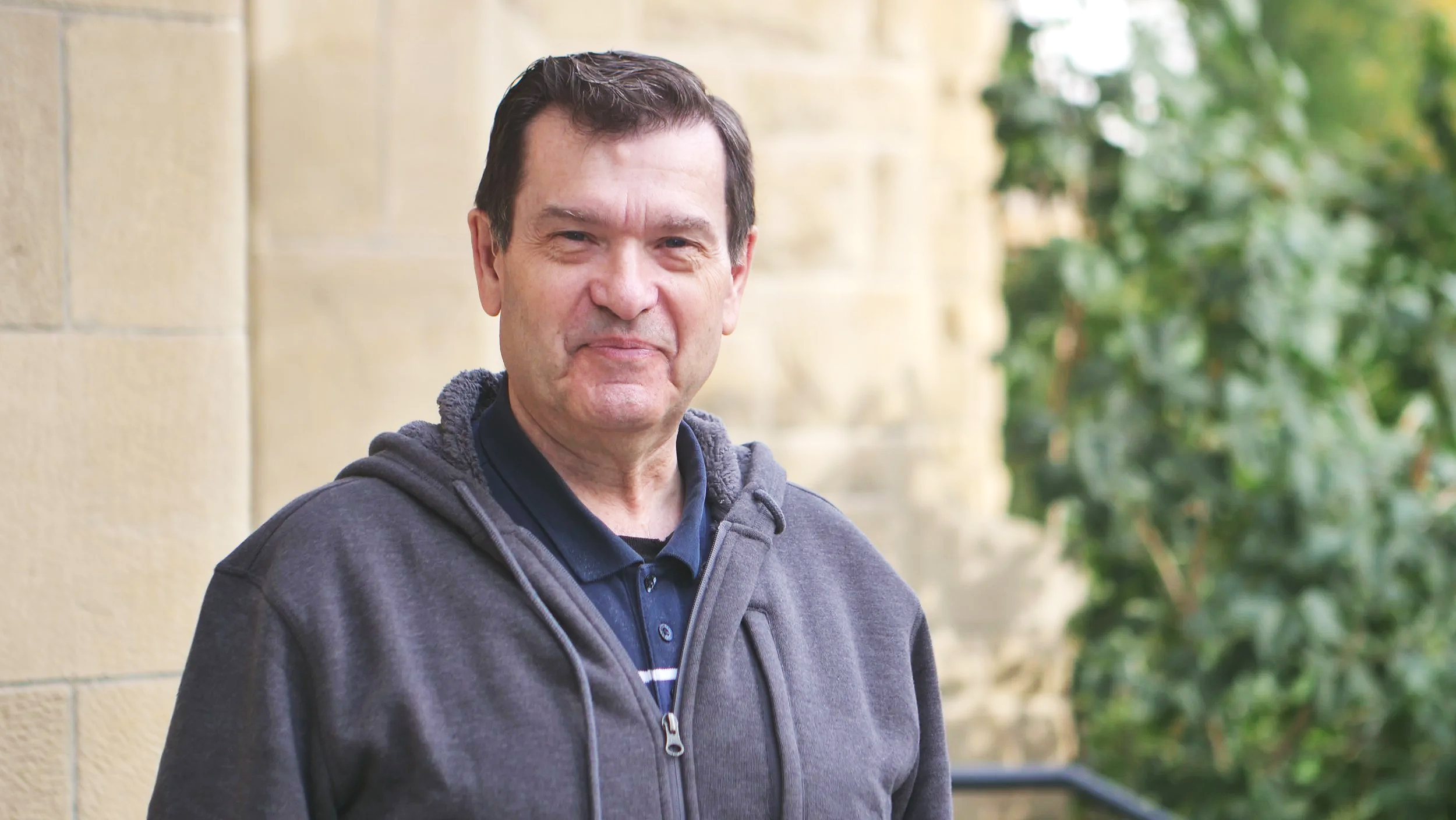 Smiling middle-aged man with dark hair, wearing a dark jacket and a polo shirt, standing outdoors near a stone building and green foliage.