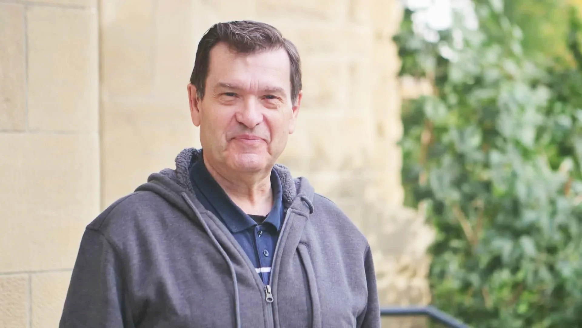 A middle-aged man with dark hair, wearing a dark blue polo shirt and a gray zip-up hoodie, standing outdoors near a stone wall and green foliage.