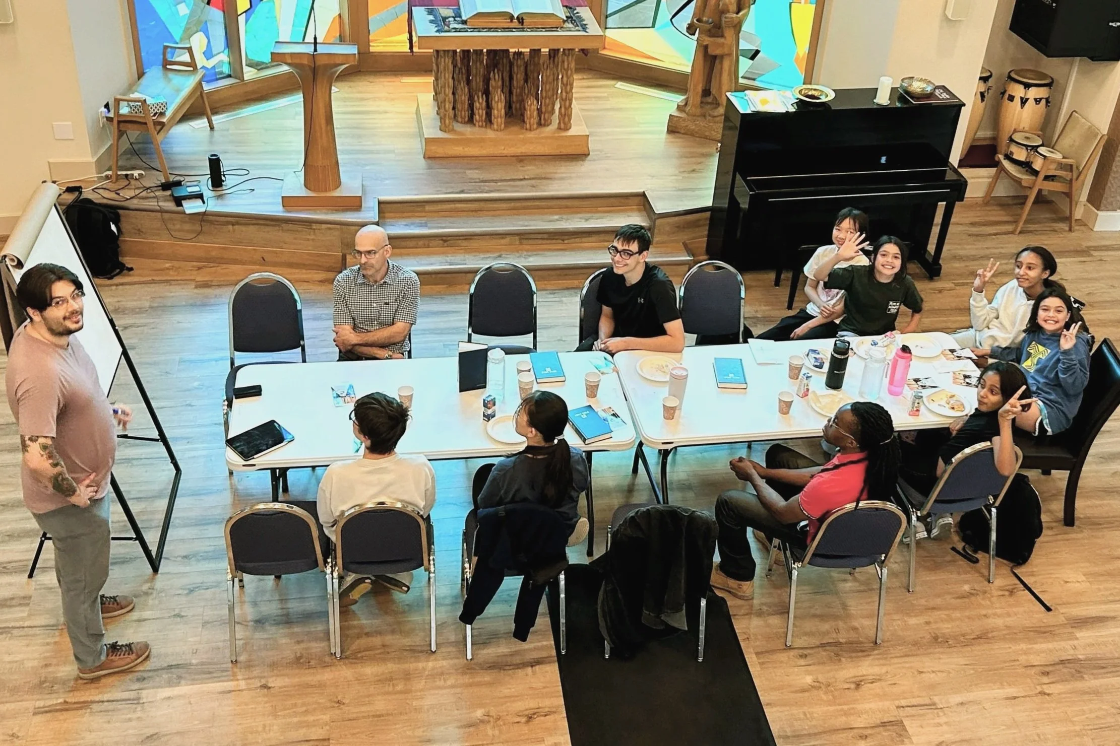 A group of youth gathered around a conference table in a room with wooden floors and colourful stained glass windows, some smiling and waving at the camera.