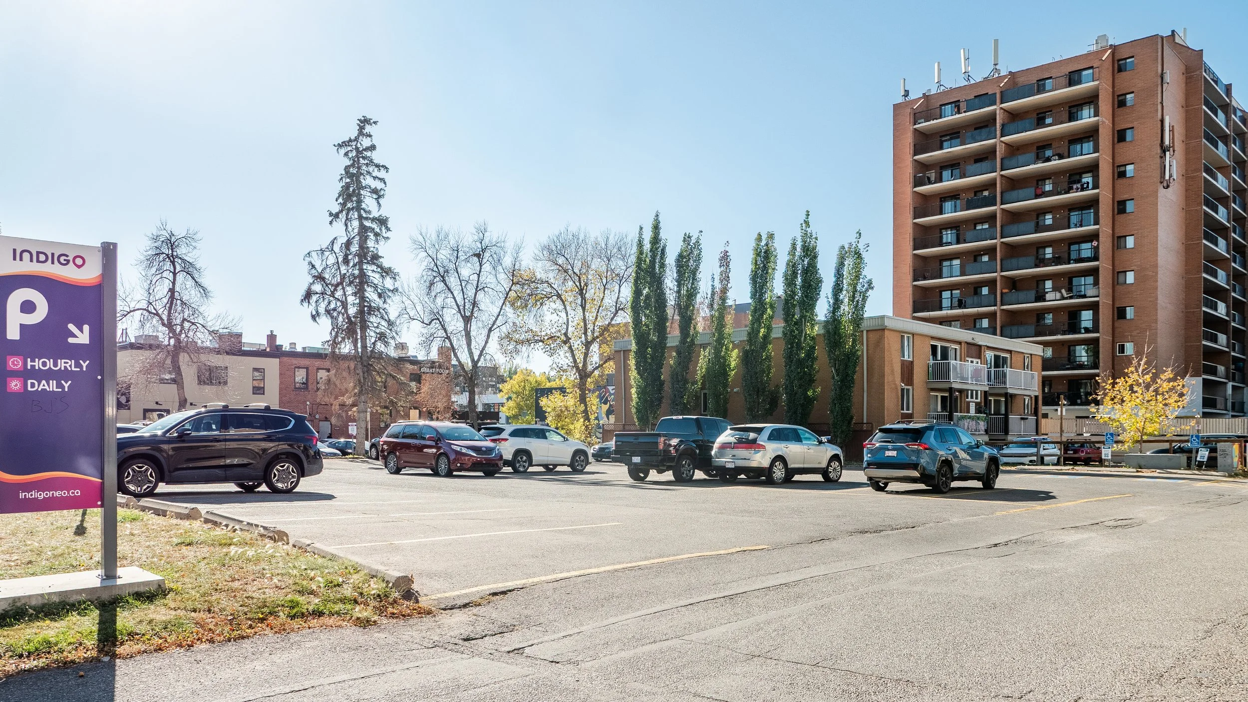 Parking lot with multiple parked cars in front of a tall apartment building and several trees, some leafless, during daytime.