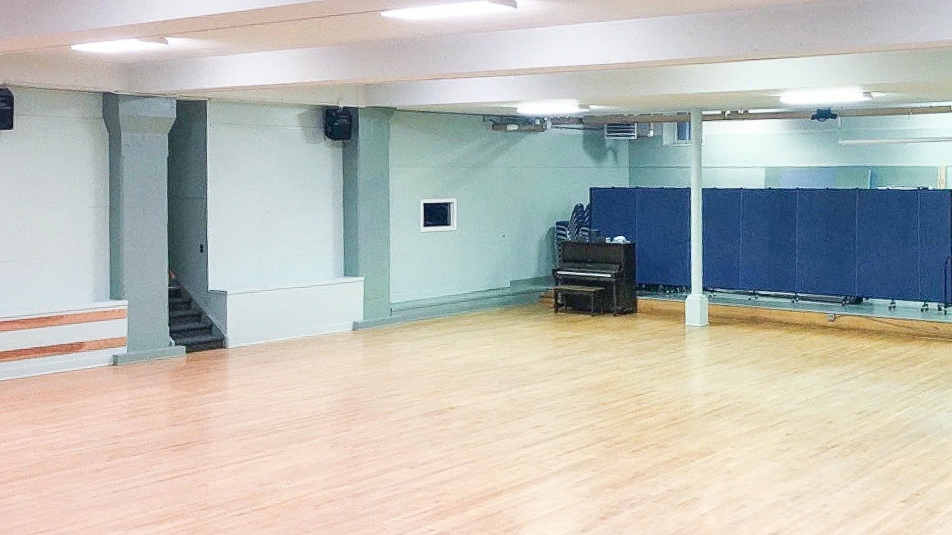Large empty dance studio or gymnasium with wooden floor, light green walls, a staircase on the left, a small window on the back wall, and blue folding partitions on the right. A piano is positioned against the back wall.