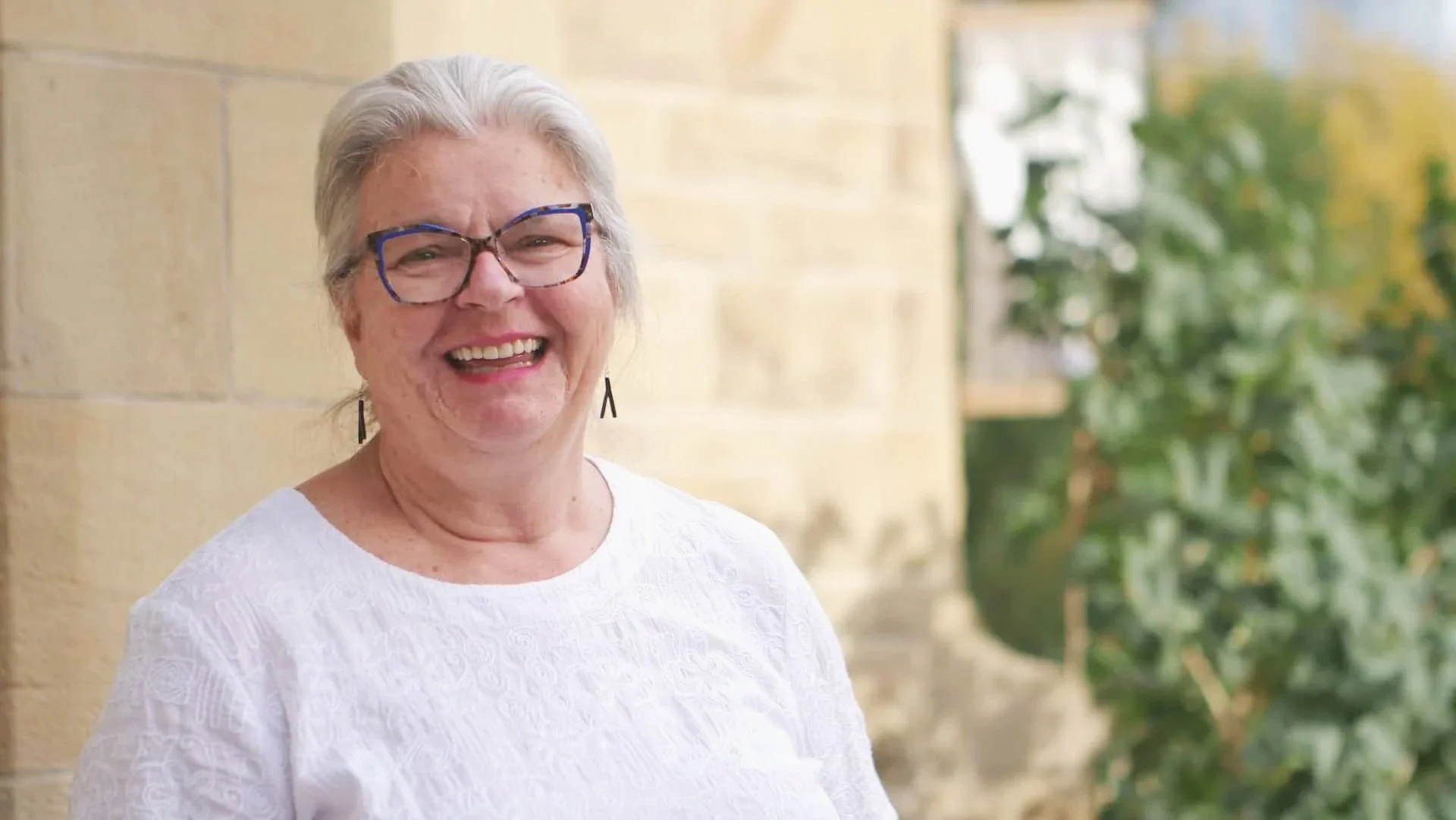 Smiling elderly woman with gray hair, glasses, earrings, wearing a white shirt, outdoors near a beige stone wall and green foliage.