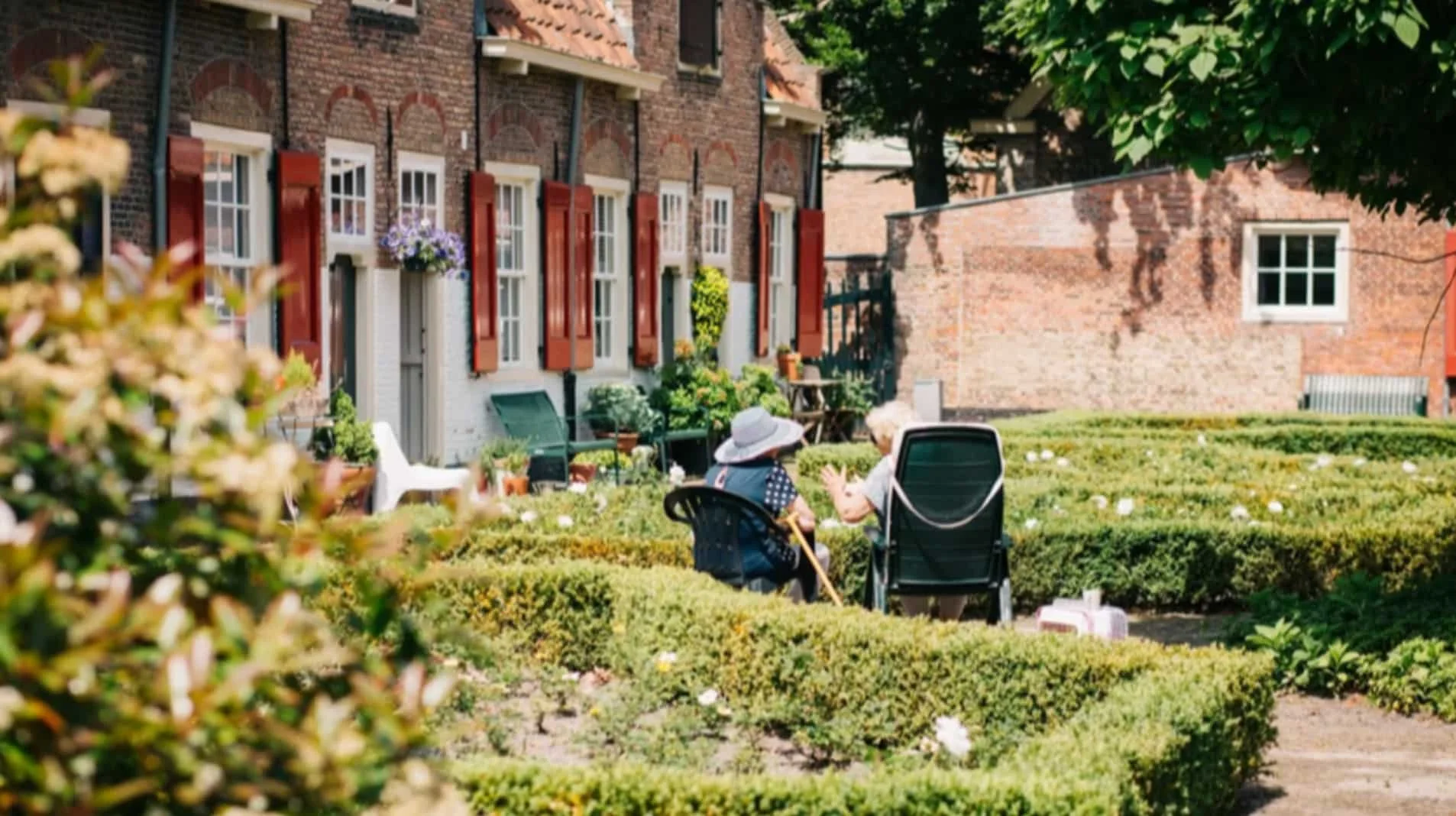 Two elderly women sitting in advance garden next to brick houses with red shutters, surrounded by neatly trimmed bushes, plants, and flowers on a sunny day.